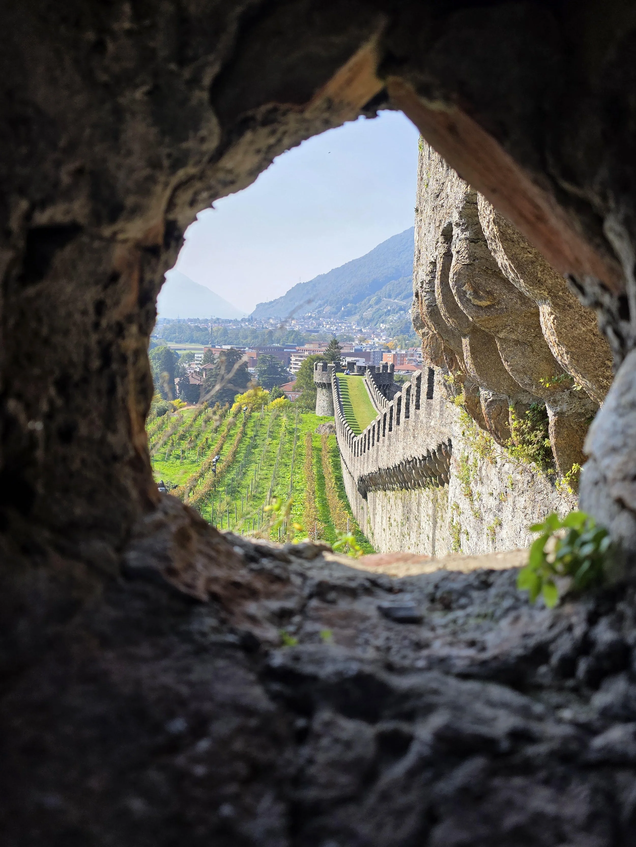 View of a scenic landscape through a volcanic rock window showing a vineyard, castle wall, town, mountains, and hazy sky in the distance.