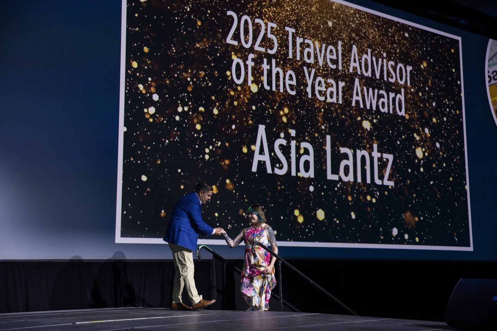 A man in a blue blazer shakes hands with a woman in a colorful dress on stage during an awards ceremony. A large screen behind them displays the text '2025 Travel Advisor of the Year Award Asia Lantz' with a glittery gold background.