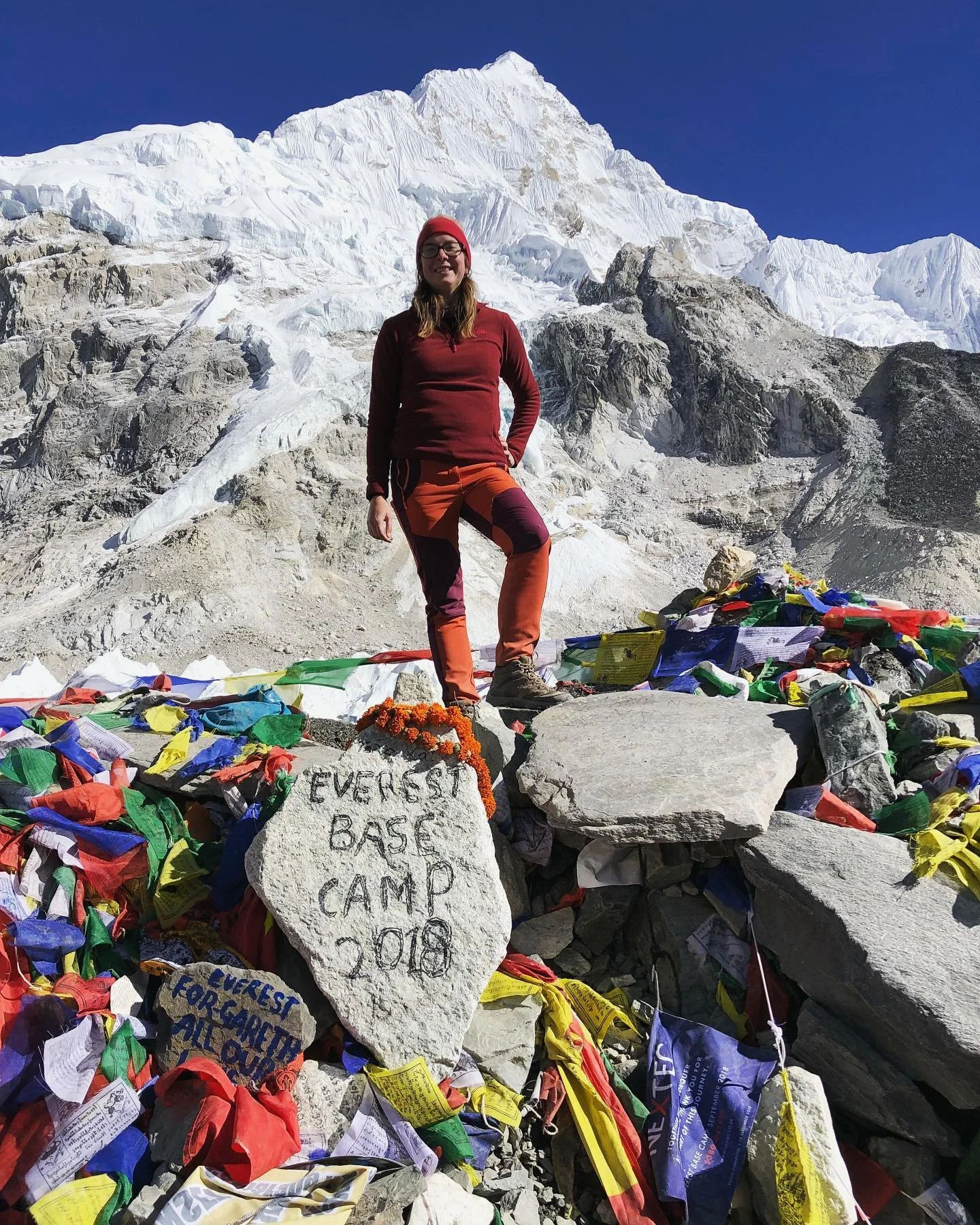 A person standing atop Everest Base Camp in 2018, with snow-covered mountains in the background and prayer flags around a large rock that has "EVEREST BASE CAMP 2018" written on it.