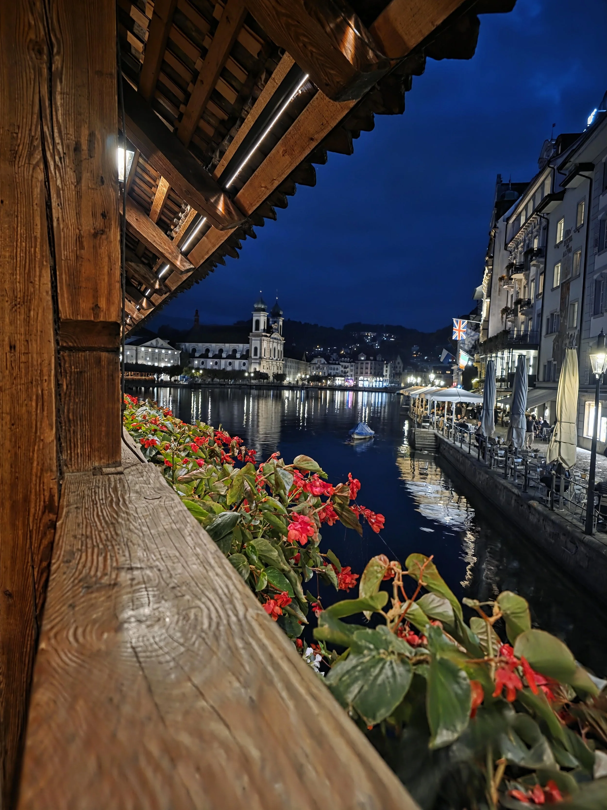 Night view of a waterfront street with buildings, flags, and outdoor dining, seen from under a wooden balcony with flowers in the foreground.