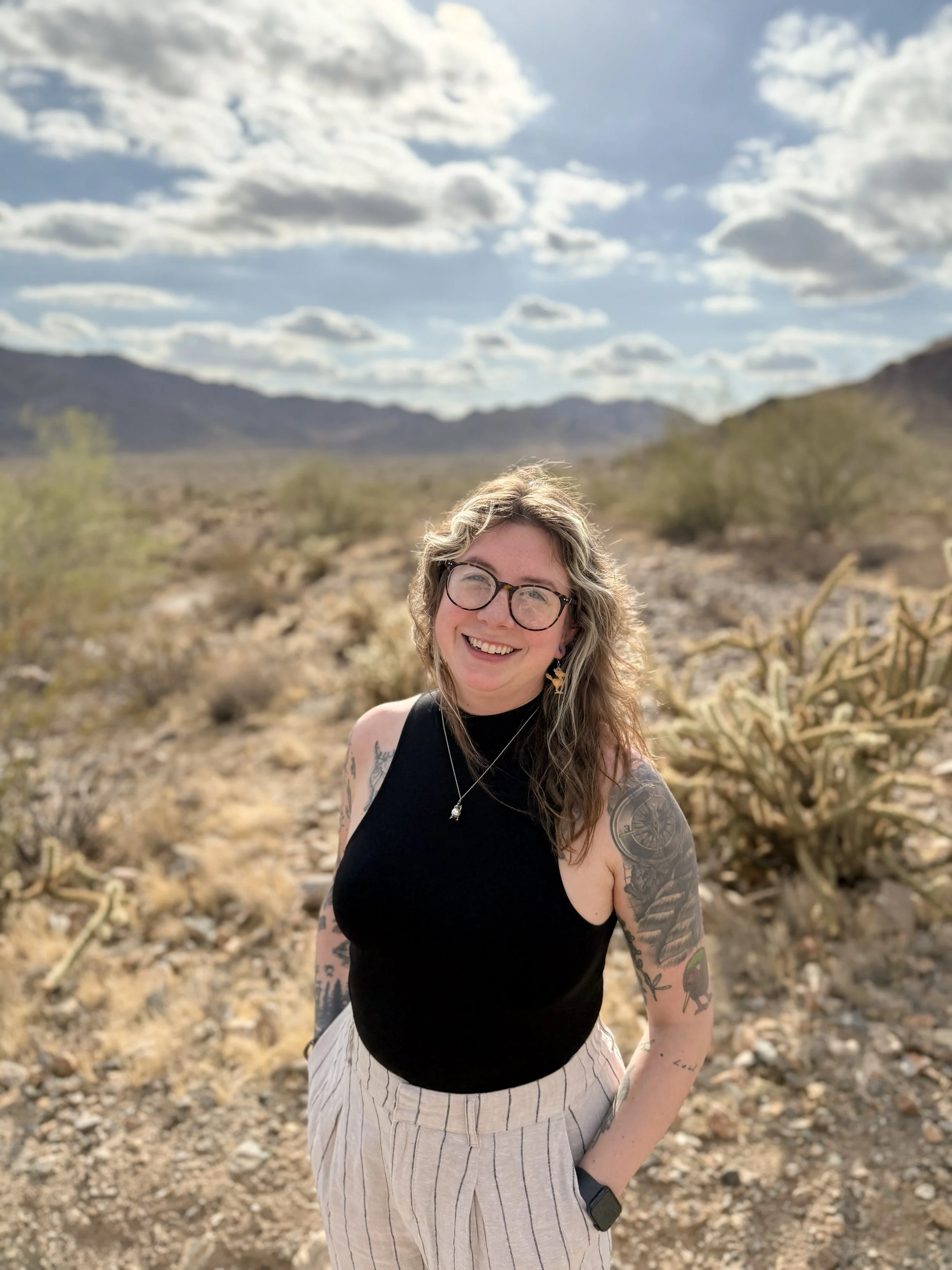 A woman with glasses and tattoos smiling outdoors in a desert landscape with mountains and a cloudy sky in the background.