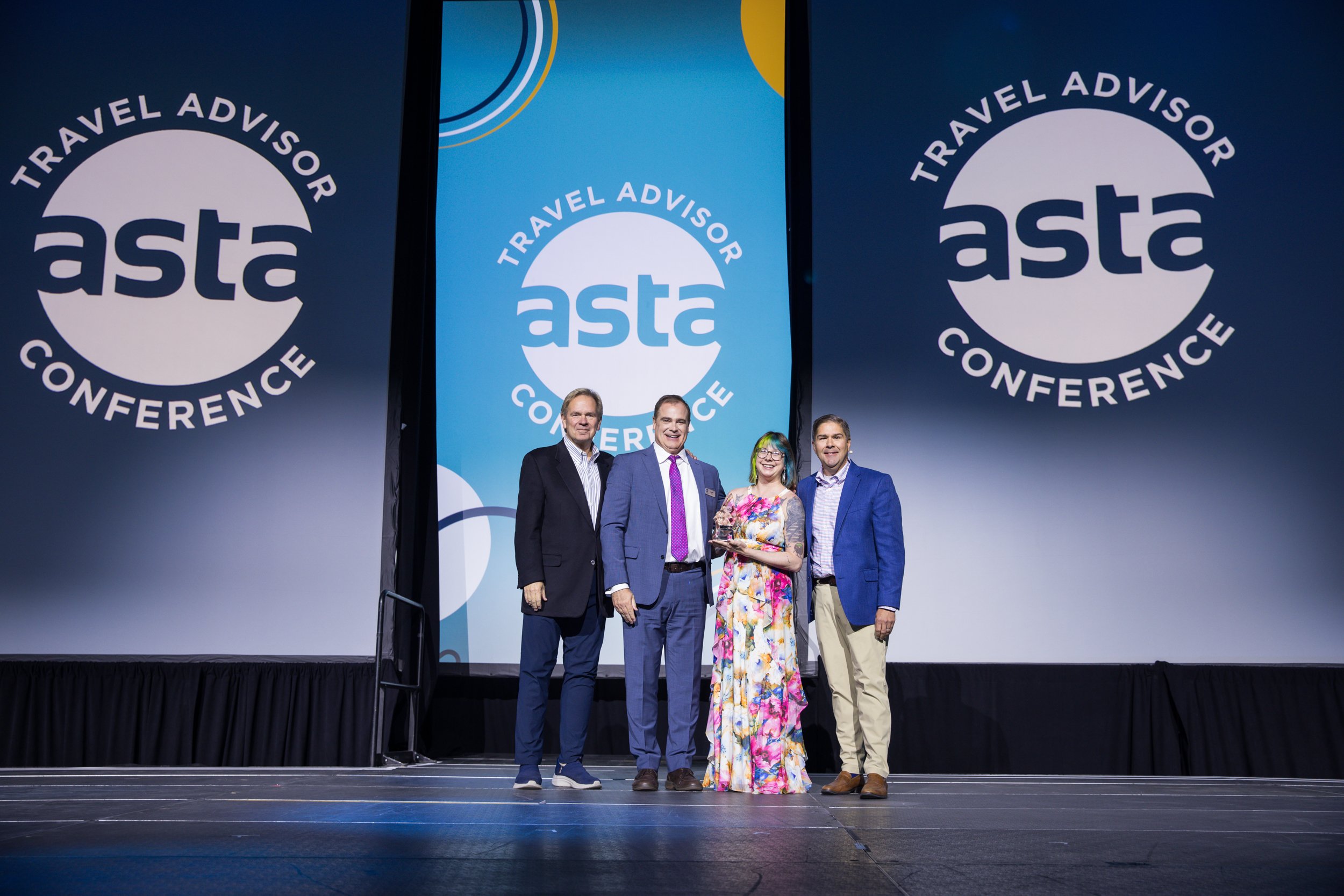 Four people standing on a stage at a conference with a large backdrop displaying the 'Travel Advisor' and 'ASTA' logos. One woman is holding an award or plaque, and all are dressed in business or semi-formal attire.