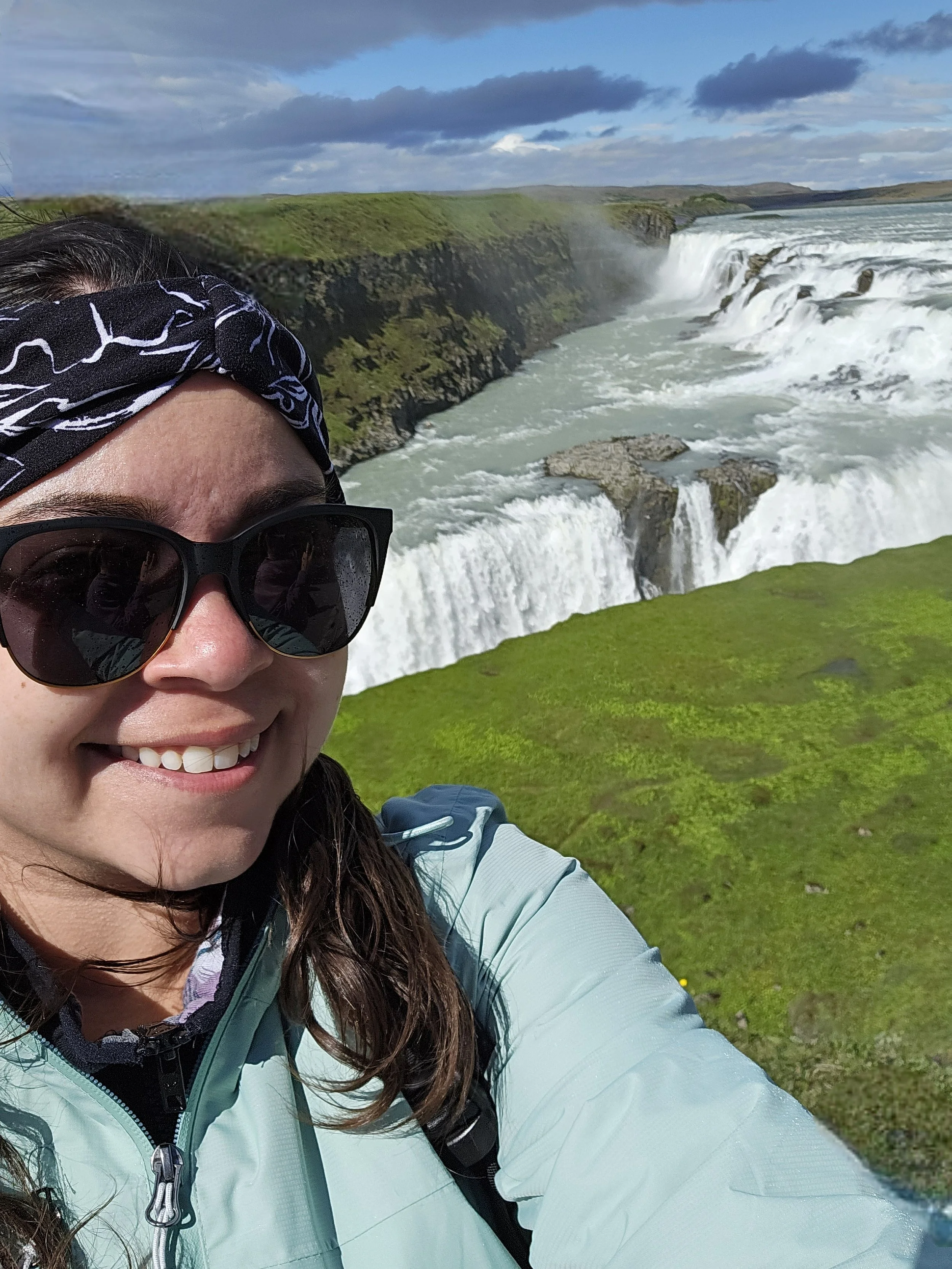 A person wearing sunglasses, a black and white headband, and a light blue jacket, taking a selfie with a waterfall and green landscape in the background.
