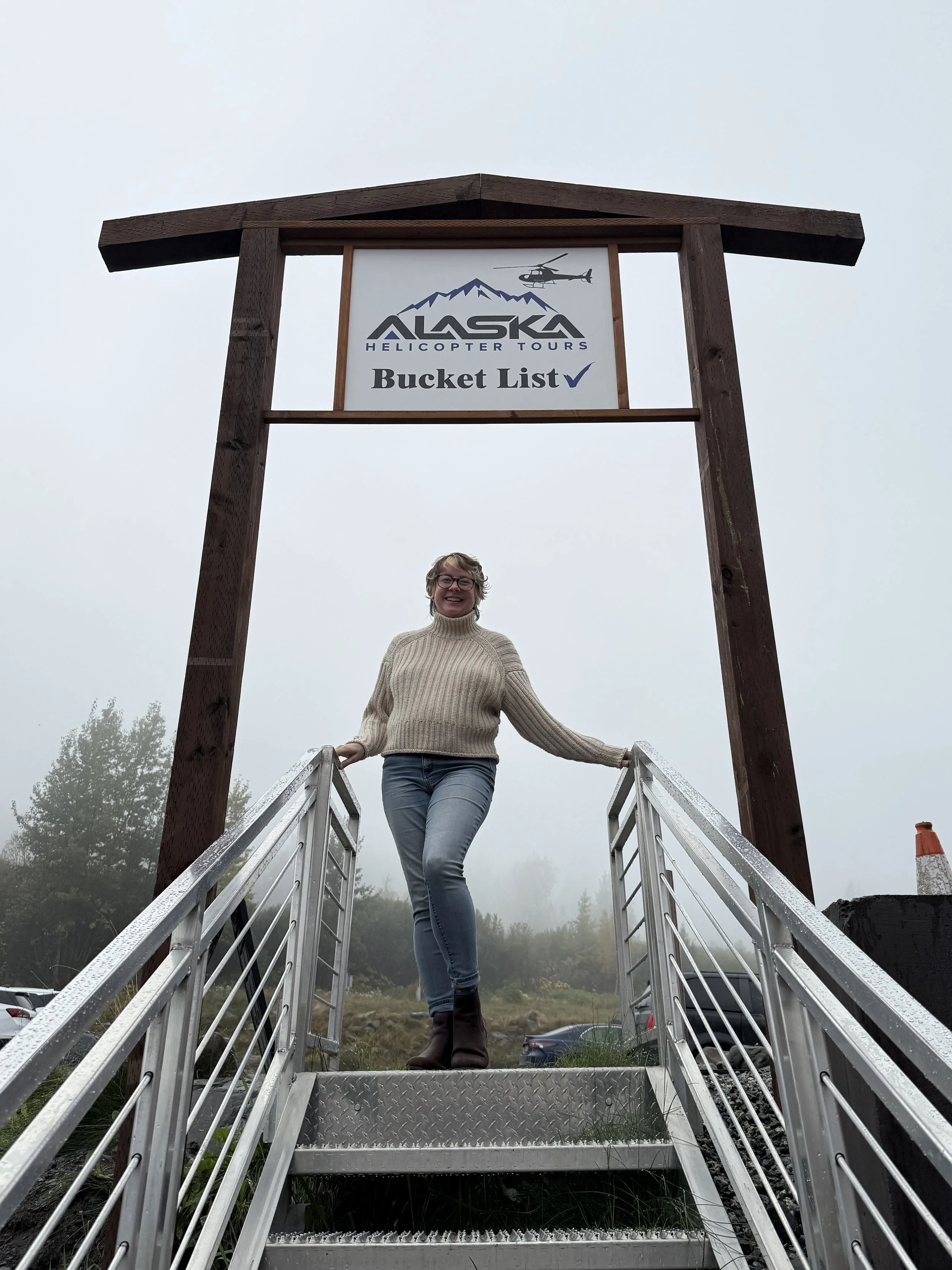 A woman standing on a metal bridge at a foggy outdoor location, smiling at the camera, wearing glasses, a beige sweater, and jeans, with a mountain and trees in the background.