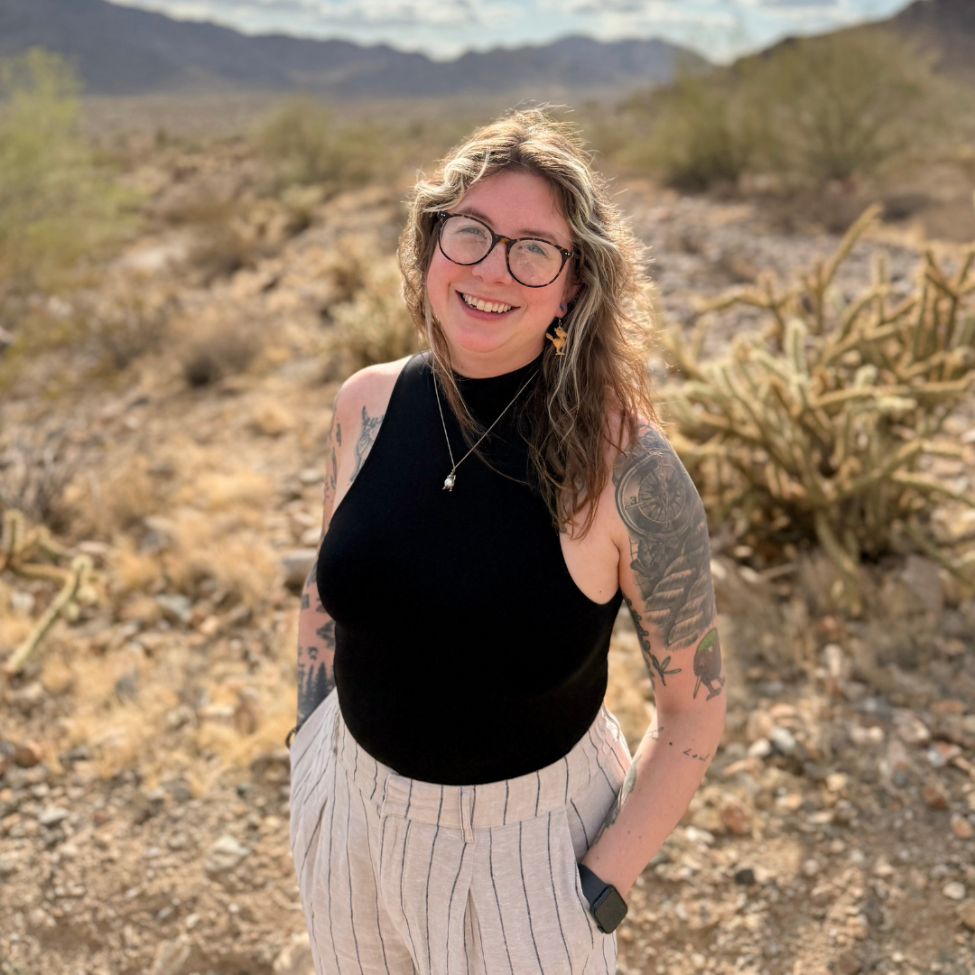 A smiling woman with glasses, curly hair, and tattoos on her arms, wearing a black sleeveless top and striped light-colored pants, standing in a desert landscape with cacti and mountains in the background.