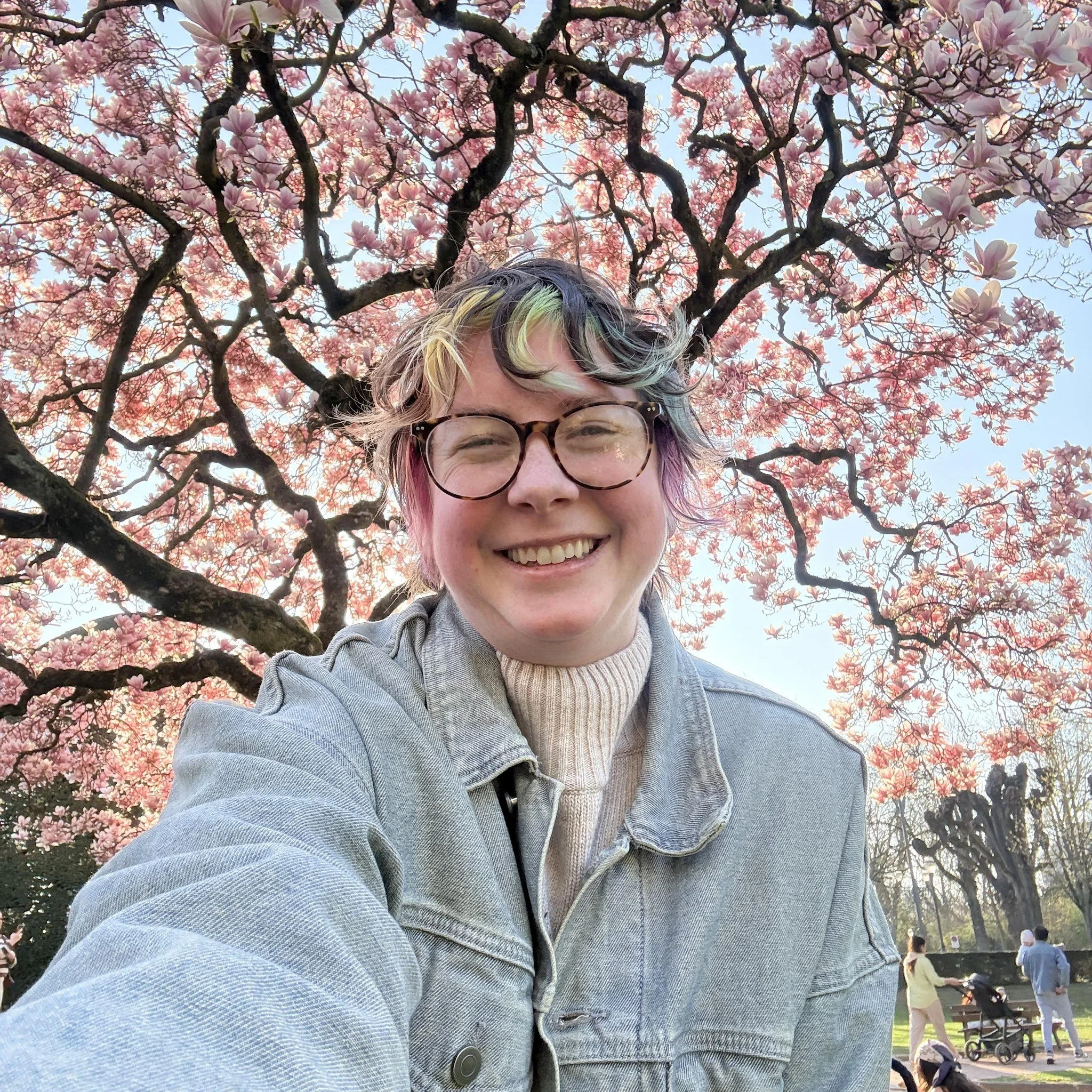 A smiling person with multicolored hair wearing glasses and a denim jacket, taking a selfie in front of a large blooming pink magnolia tree.