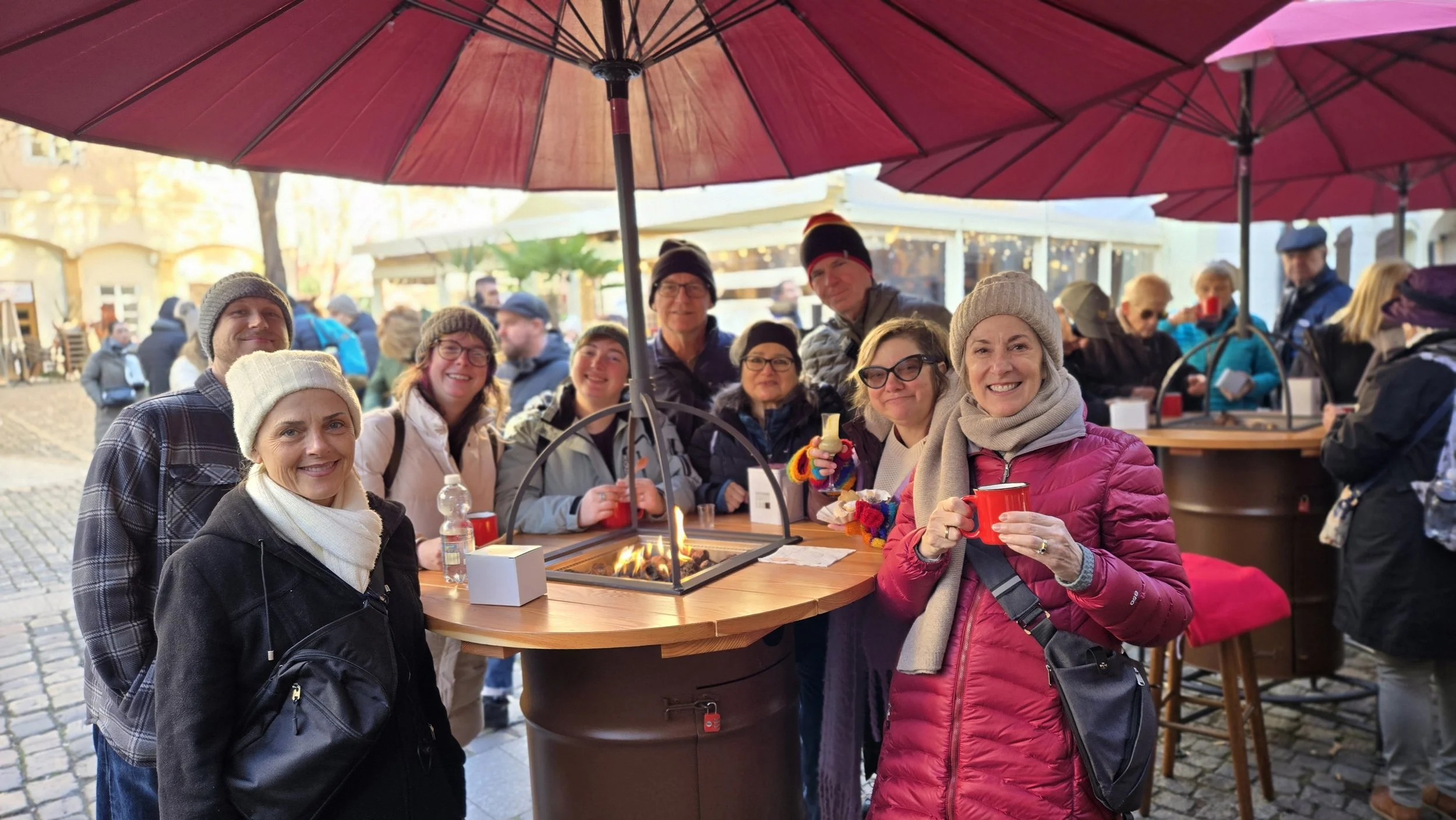 A group of people gathered around a round table under large red umbrellas outdoors during daytime, enjoying food and drinks with a fire pit in the center, smiling and wearing warm clothes.