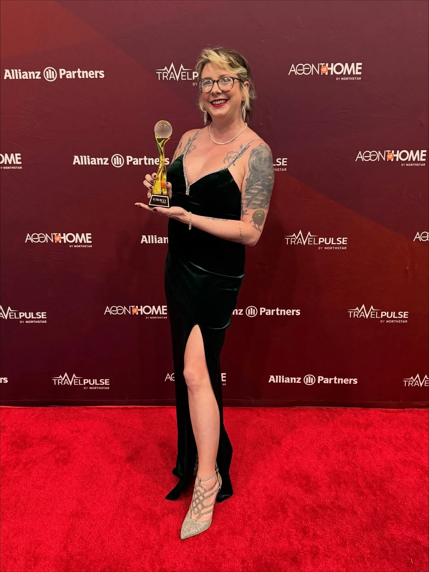 A woman with tattoos wearing a black dress with a high slit, holding a trophy, posing on a red carpet with a red backdrop that has logos including Allianz Partners, TravelPulse, and AGENTHOME.