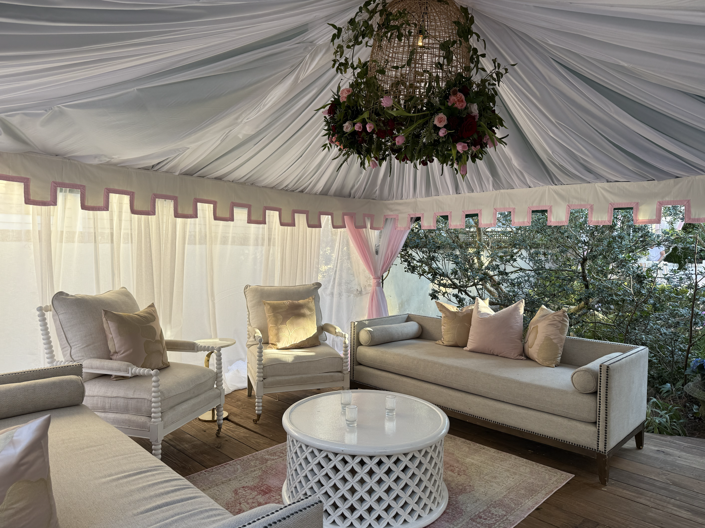 Elegant outdoor seating area under a decorated tent with white fabric ceiling, pink trimming, a floral chandelier, beige sofas and armchairs with pastel cushions, a white round coffee table, and lush greenery outside.