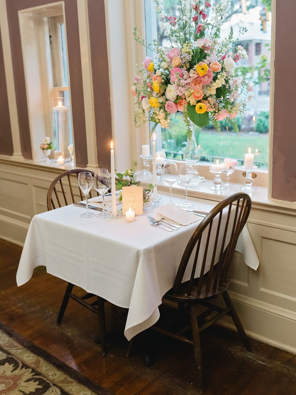 A romantic table setting by a large window with a tall, colorful flower arrangement, lit candles, wine glasses, and a white tablecloth.