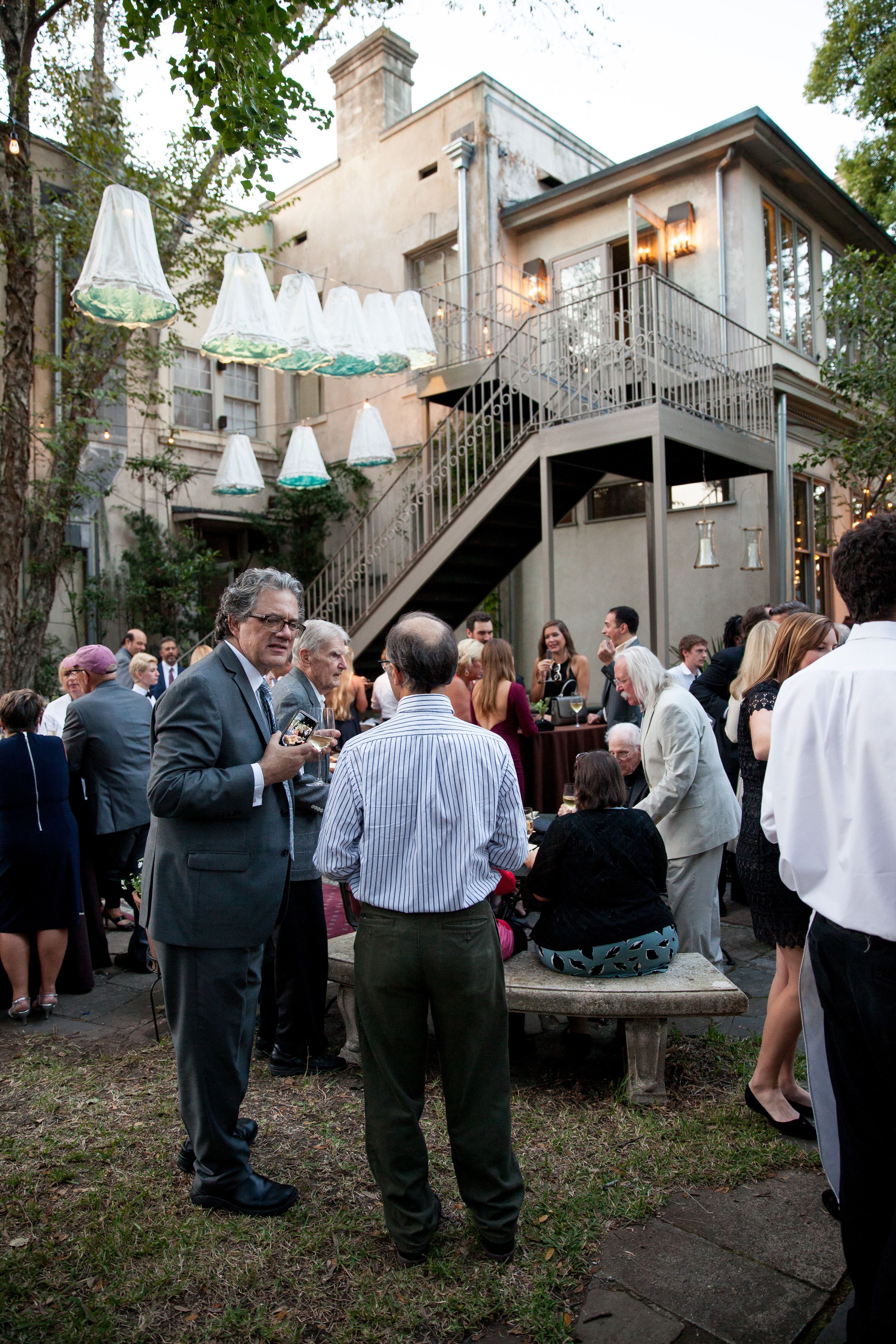 People socializing outdoors at a gathering in a backyard with a two-story house, string lights, and hanging lamps.