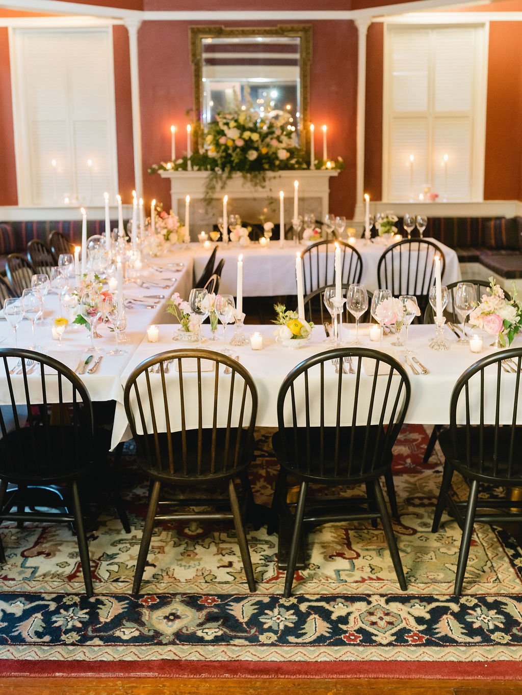 Elegant dining room set for a celebration with white tablecloths, tall candles, floral centerpieces, and glassware, with a fire place and mirror decorated with flowers and candles in the background.
