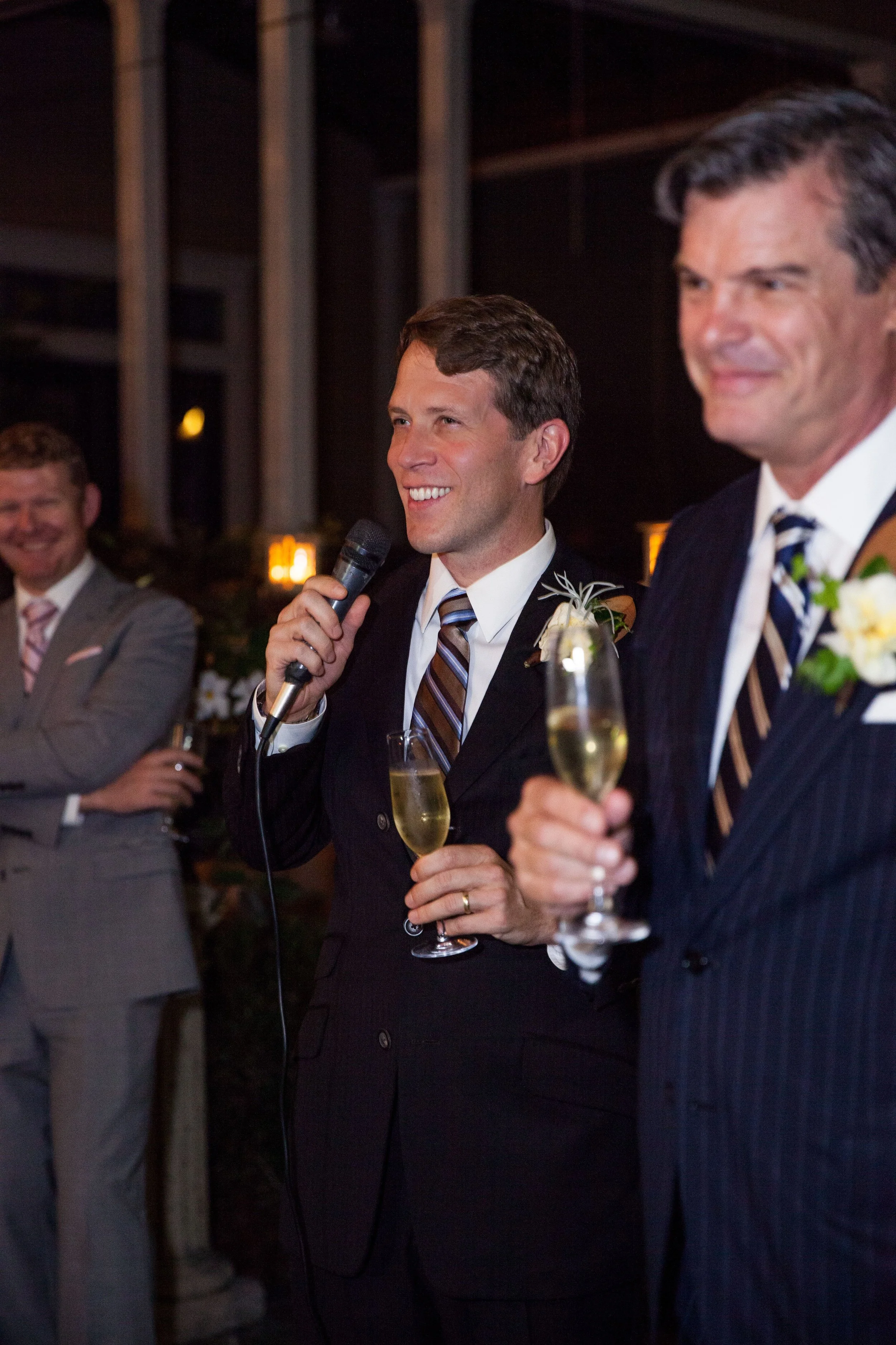 A man in a suit holding a microphone and a champagne glass, speaking at a celebration with other men in suits holding drinks.