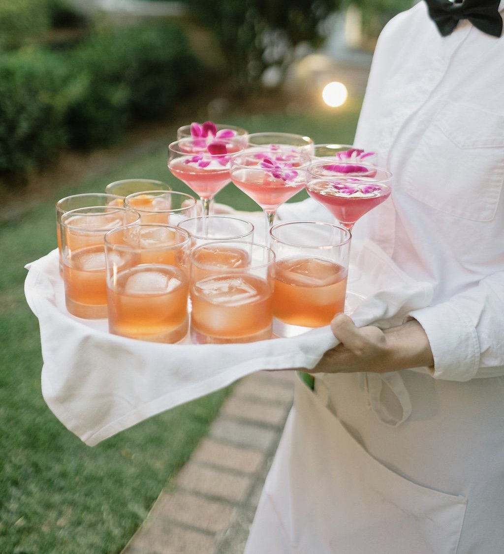A person wearing a white shirt and black bow tie holding a tray with pink and orange drinks garnished with edible flowers, outdoors at sunset.