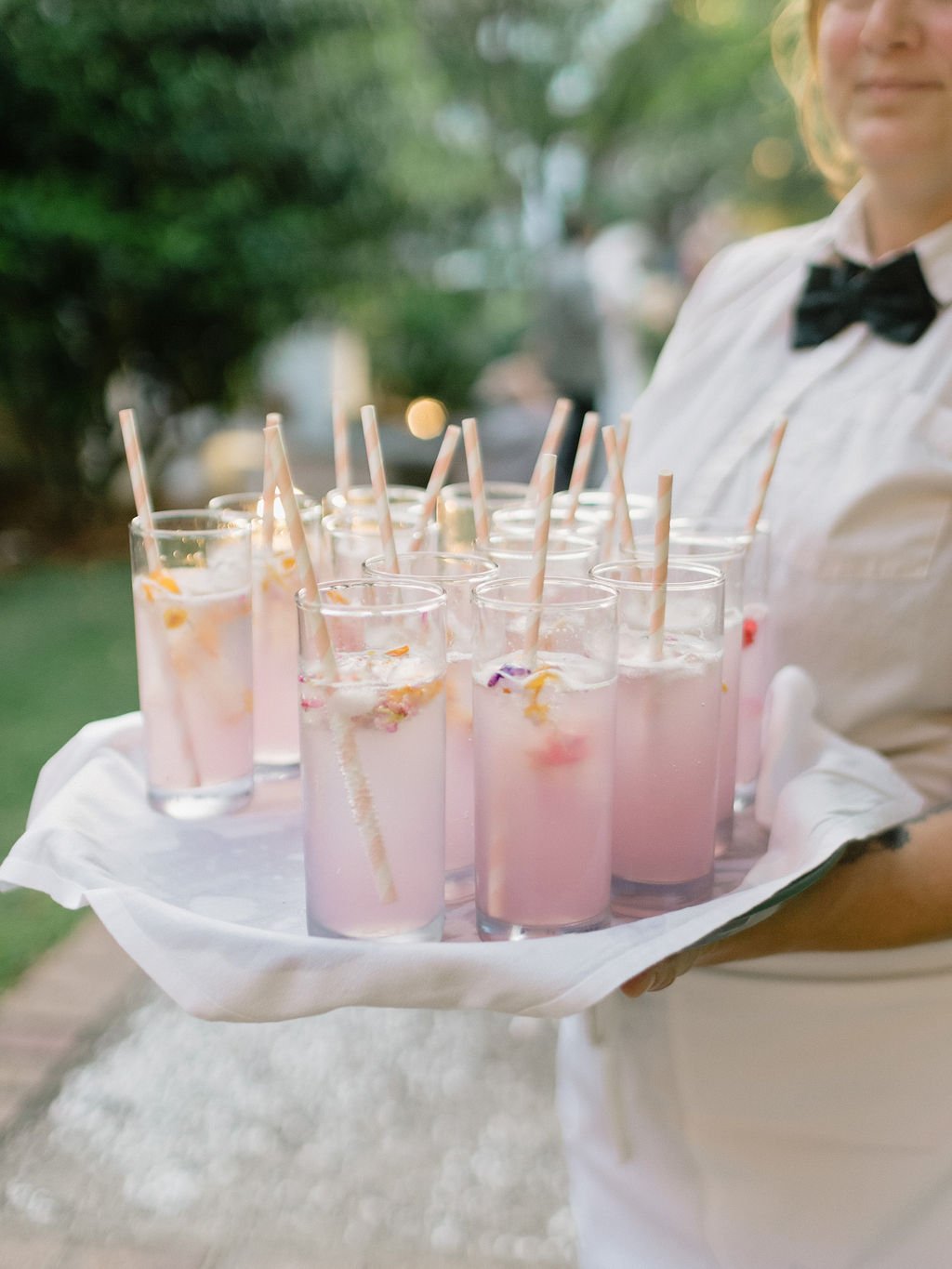 A server holding a tray with multiple pink beverages in tall glasses, each with a striped straw, set outdoors with greenery in the background.