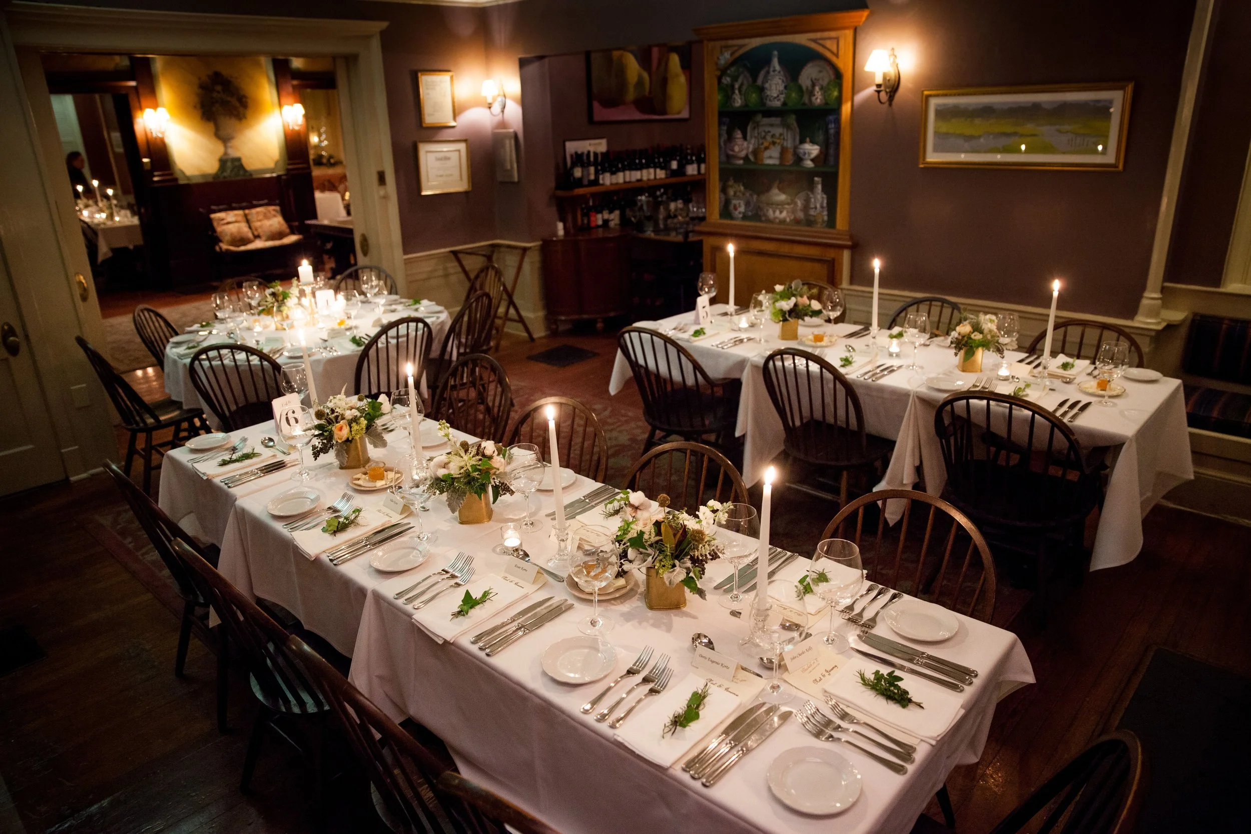 Elegant dining room with four long tables set for a formal event, decorated with white tablecloths, floral centerpieces, tall candles, and place settings with plates, glasses, and silverware, in a warmly lit room.