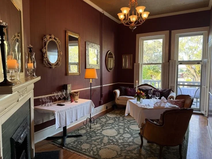 Elegant dining room with deep red walls, a chandelier, and large windows with white shutters. The room features a sofa, a table with a floral centerpiece, and a side table with glasses and candles.