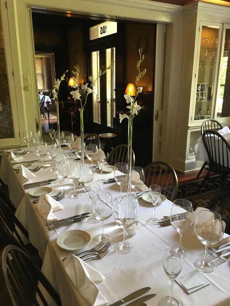 A dining table set for a formal meal with white tablecloth, multiple wine glasses, plates, silverware, and napkins folded into triangles, decorated with tall glass vases holding white lilies with long stems and green leaves.