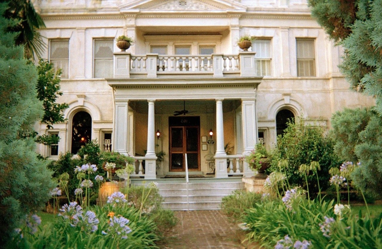Front view of a large, historic-style house with a porch and columns, surrounded by lush plants and flowers.