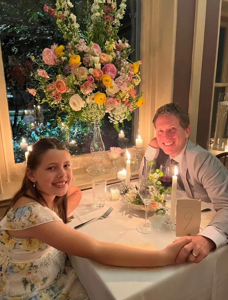 A young girl and an older man sitting at a decorated dinner table with candles and a large floral centerpiece, smiling and holding hands. The setting is elegant with a window and candles in the background.
