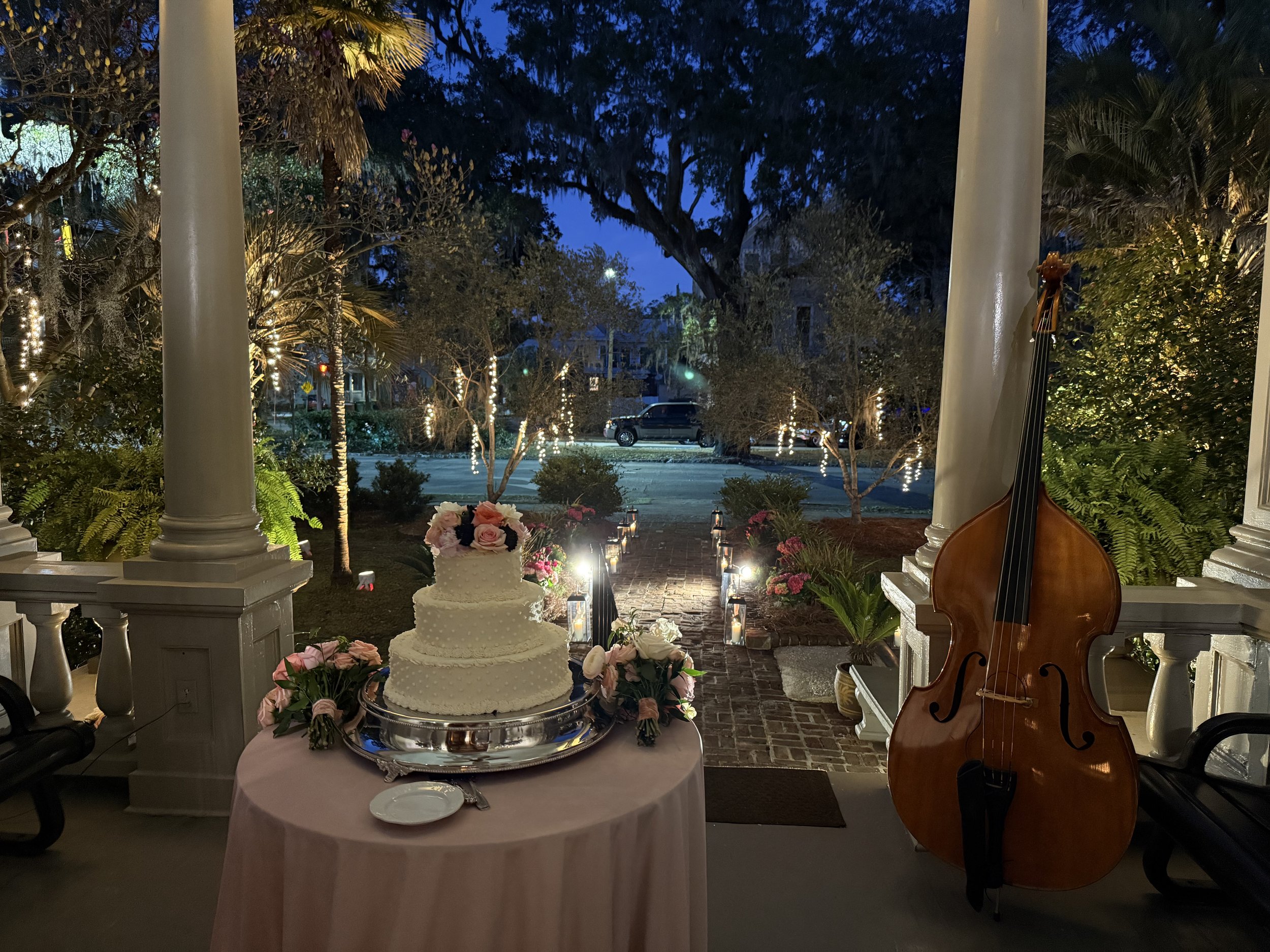 A decorated outdoor porch at dusk with a wedding cake on a pink table, surrounded by pink roses, a double bass leaning on the porch railing, and a garden path illuminated by candles and string lights.