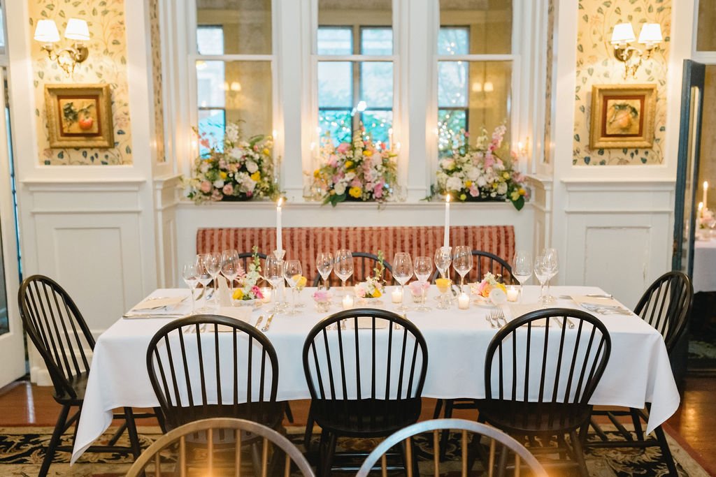 A decorated dining table with white tablecloth, wine glasses, candles, and floral centerpieces set for a meal in a room with floral wallpaper and wall lamps.