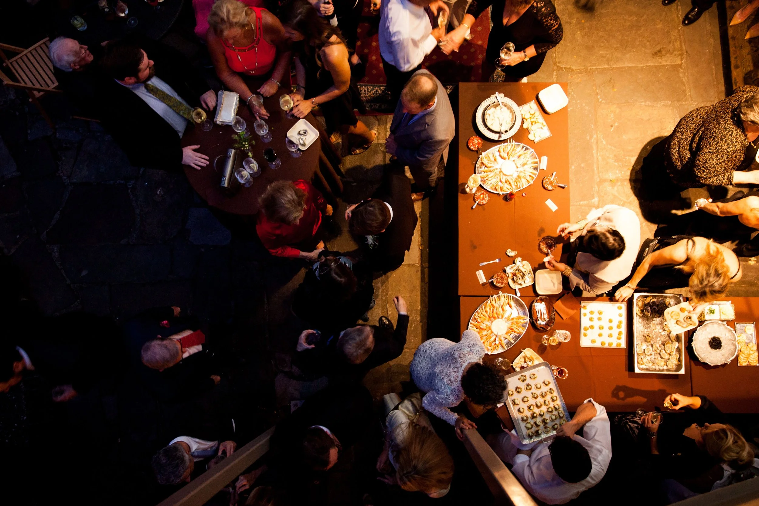Overhead view of a social gathering with people standing and sitting around a buffet table filled with various appetizers and desserts, illuminated by warm lighting.