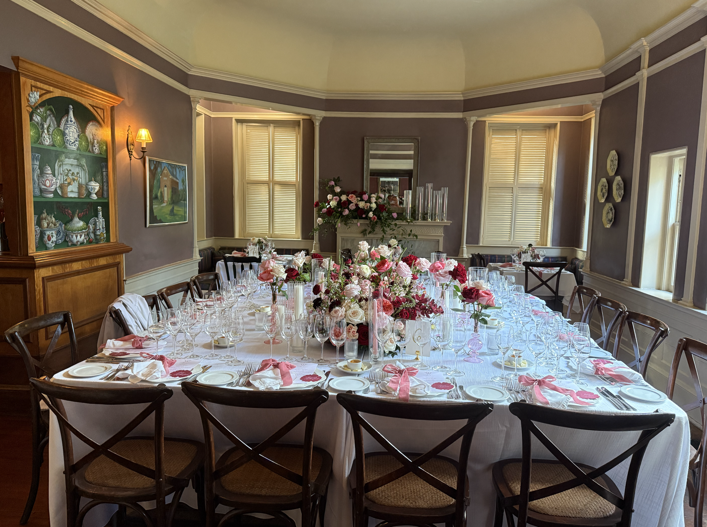 Elegant dining room set for a formal event with a large white tablecloth, floral centerpieces, and place settings with wine glasses and pink napkins. The room has purple walls, multiple windows with shutters, and decorative items on the walls.