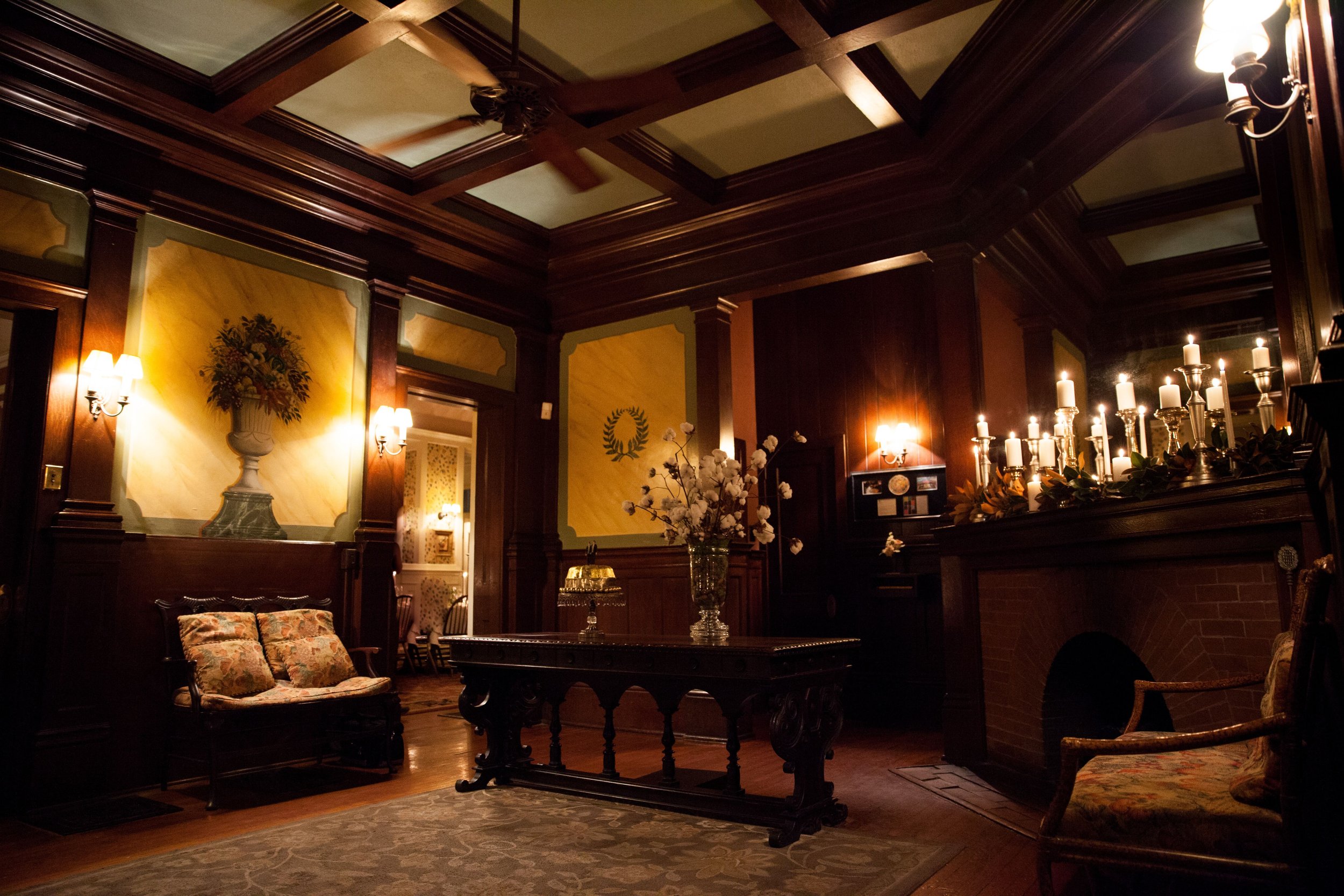 A dimly lit, elegant living room with dark wood paneling, a fireplace with candles, floral arrangements, and vintage furniture.