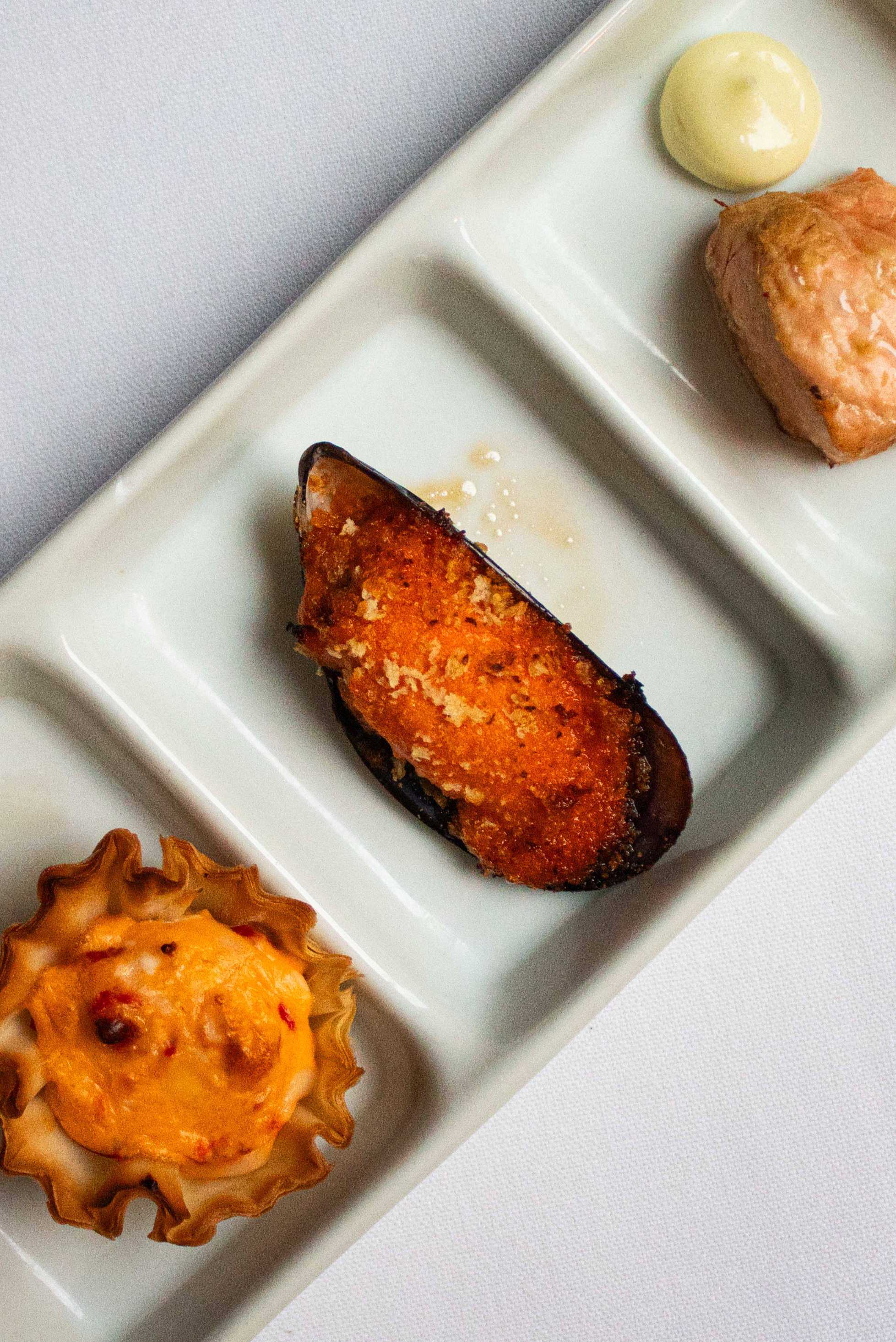 Three dishes on white ceramic serving trays, including a stuffed mushroom with cheese, a stuffed eggplant with breading, and a meatball with sauce and a dollop of white sauce.