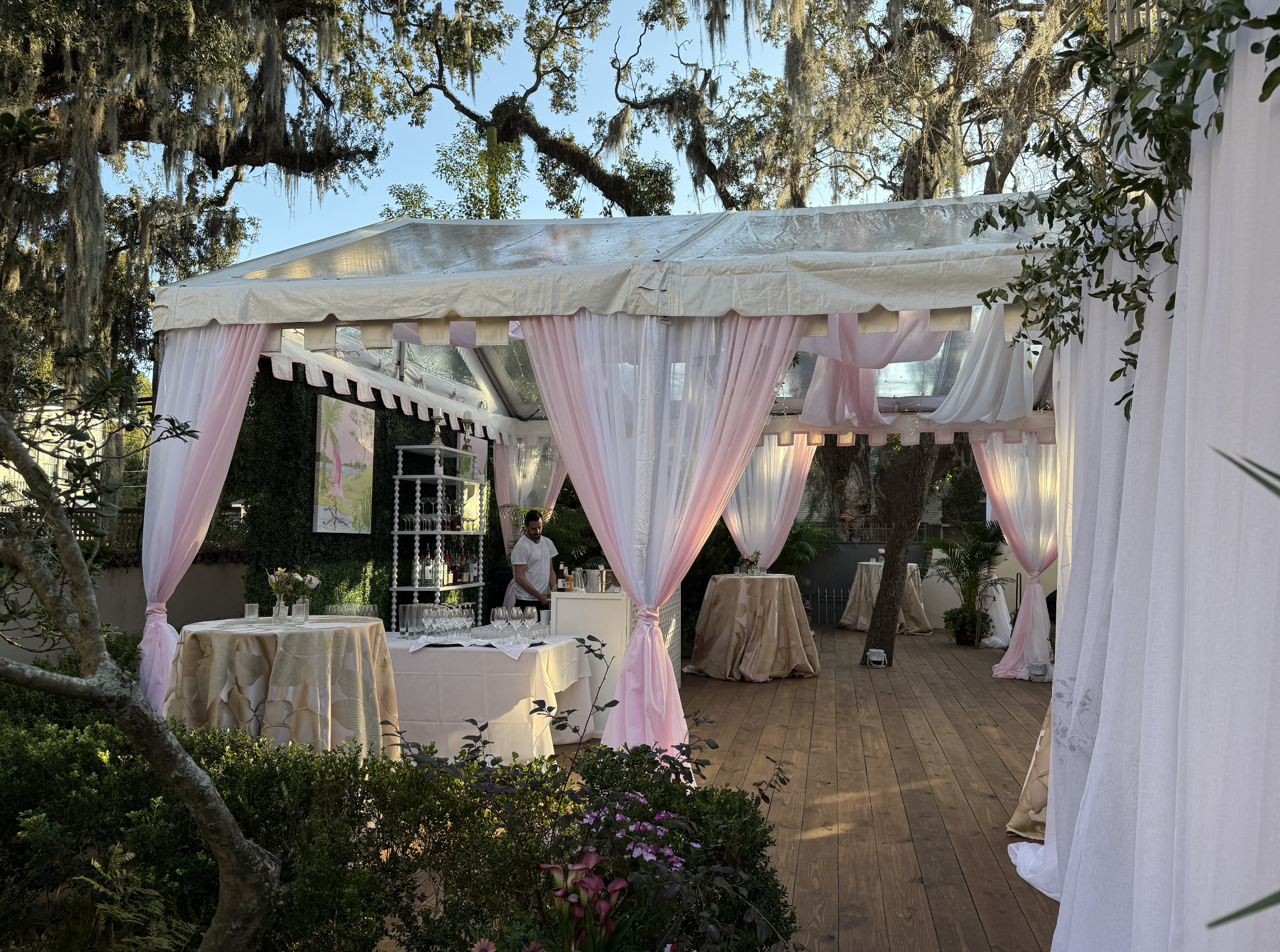 Outdoor event setup with a white canopy tent decorated with pink and white drapes, tables with beige tablecloths, and a bartender preparing drinks.