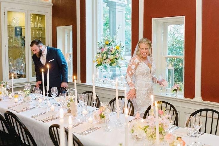Bride in a lace wedding dress smiling and holding a glass, standing near a decorated banquet table with lit candles for a wedding celebration.