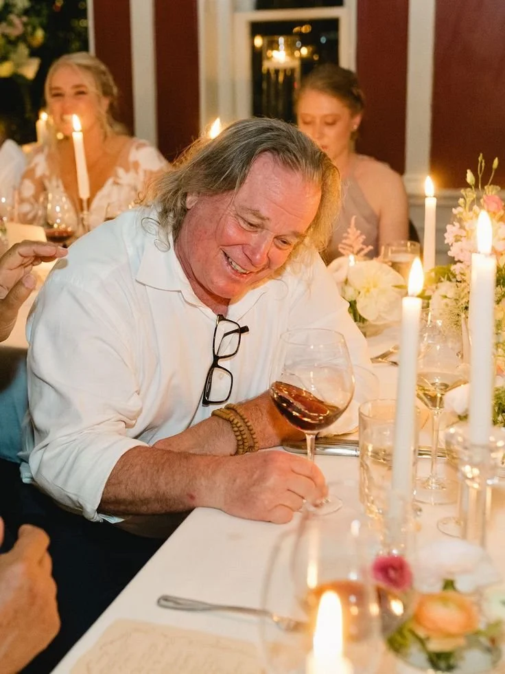 A man with long hair, wearing a white shirt and glasses hanging from his collar, is sitting at a dining table, smiling, and holding a glass of red wine. The table is decorated with lit candles, flowers, and glassware. In the background, women are als