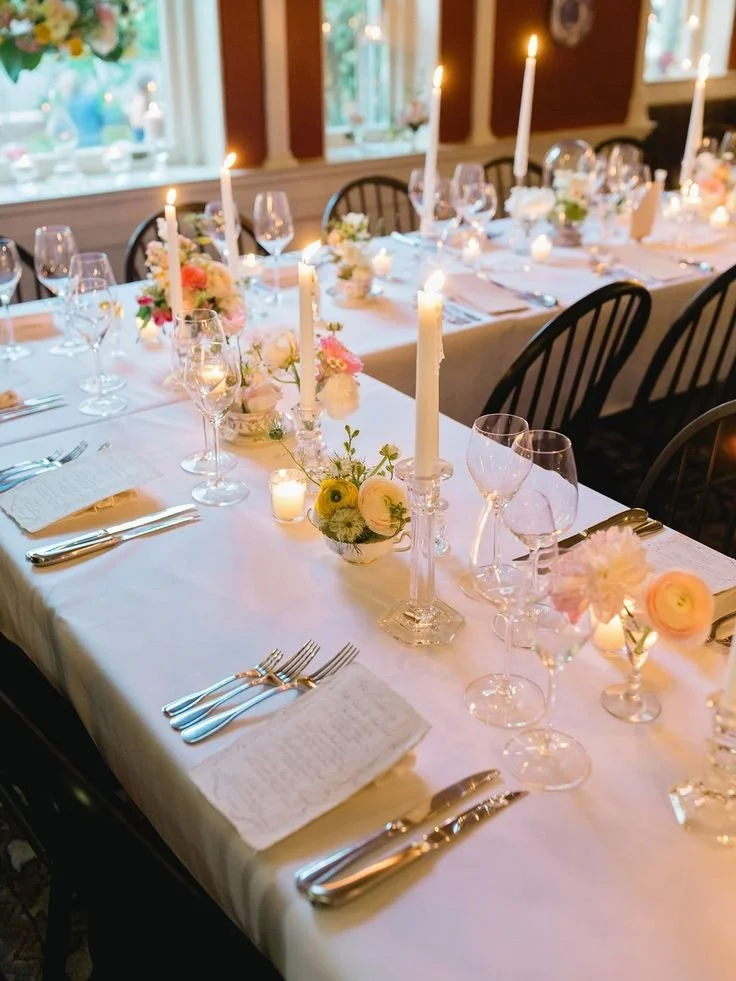 Elegant dining table set for a celebration, with white tablecloths, floral centerpieces, tall white candles, and neatly arranged glassware and silverware.
