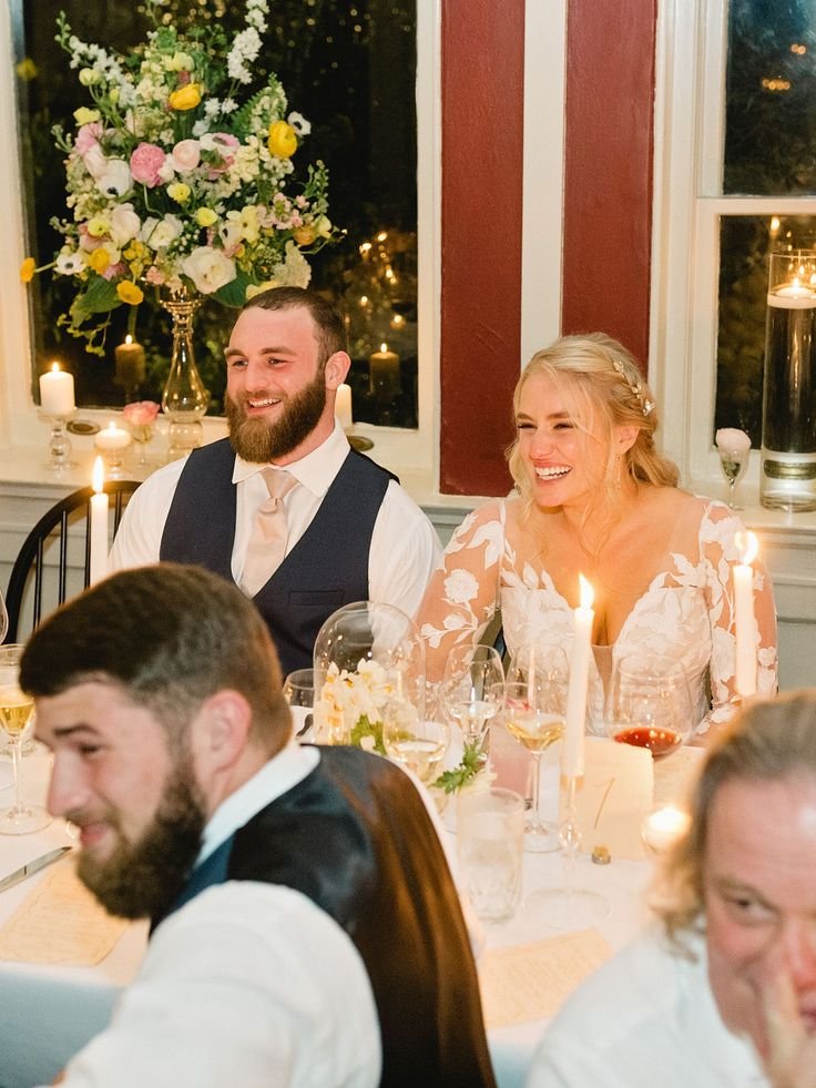 Happy bride and groom sitting at their wedding reception table, surrounded by friends, with floral arrangements and candles in the background.