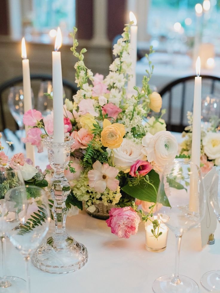 Elegant table centerpiece with pastel pink, white, and yellow flowers, surrounded by lit white candles in crystal holders, set on a white tablecloth with glasses and tableware in the background.