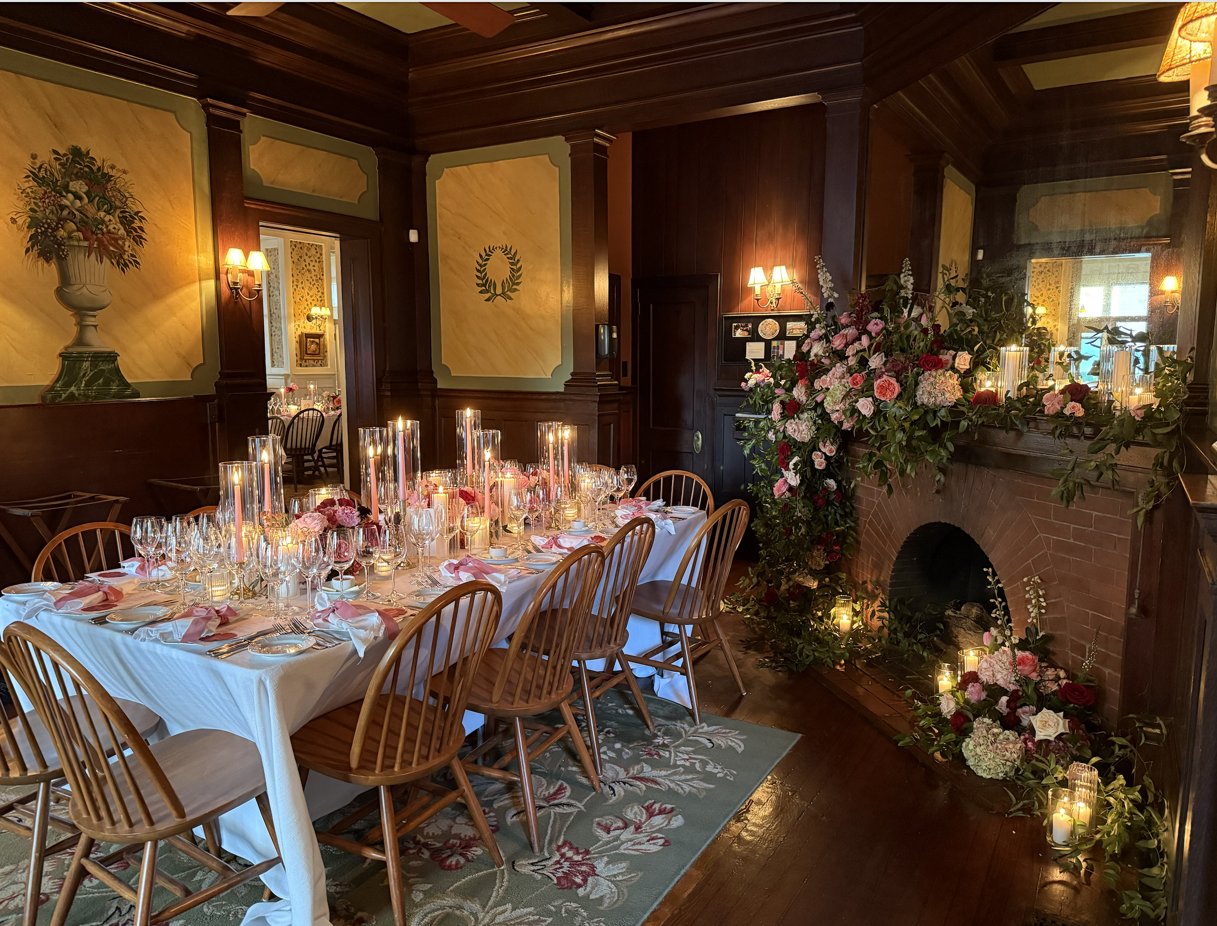 Elegant dining room decorated for a special occasion with a long table, pink and red floral centerpieces, tall candles, and a floral arrangement near a brick fireplace.