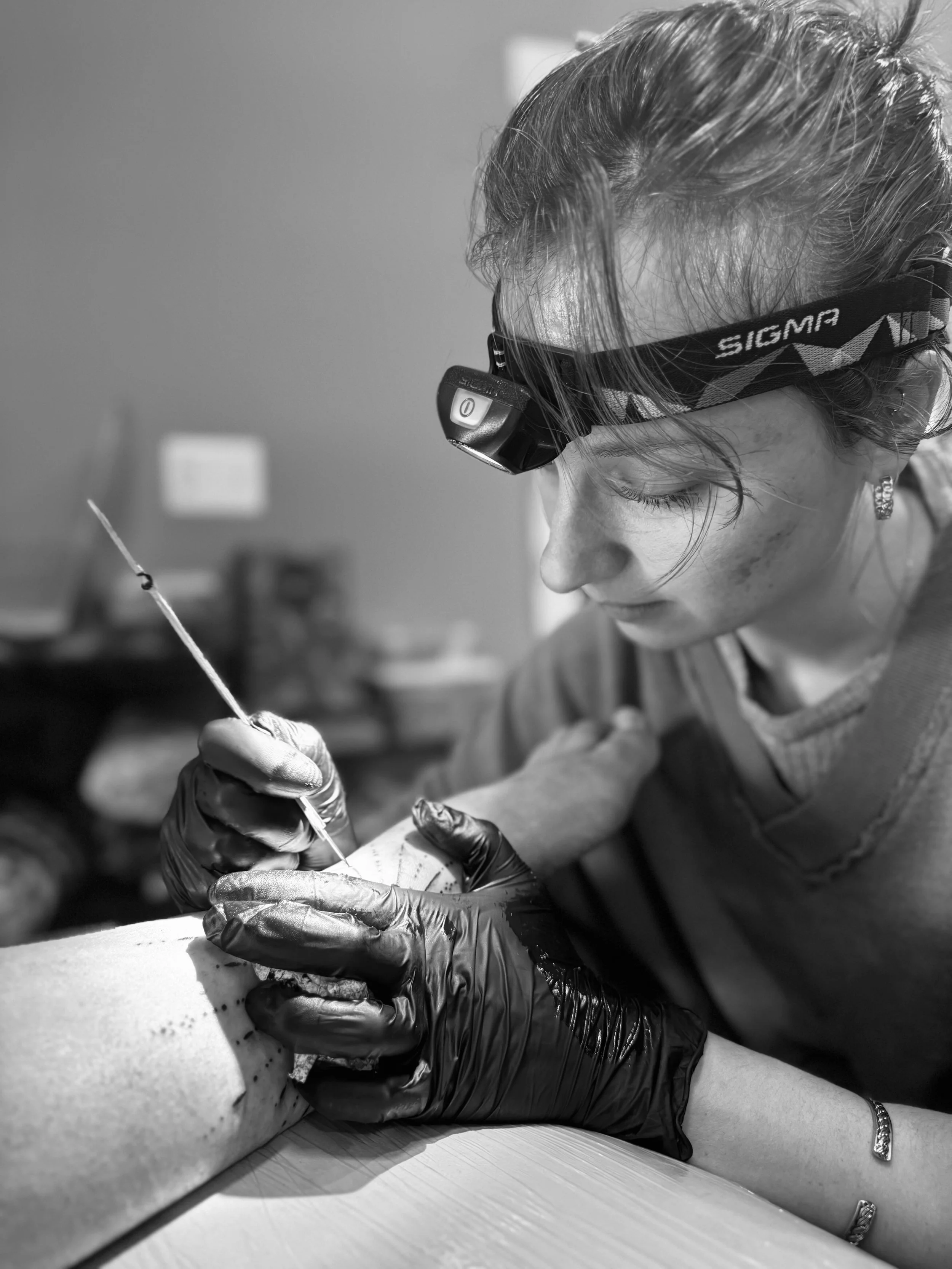 A woman wearing a headlamp and black gloves is tattooing a person's arm with a tattoo needle.