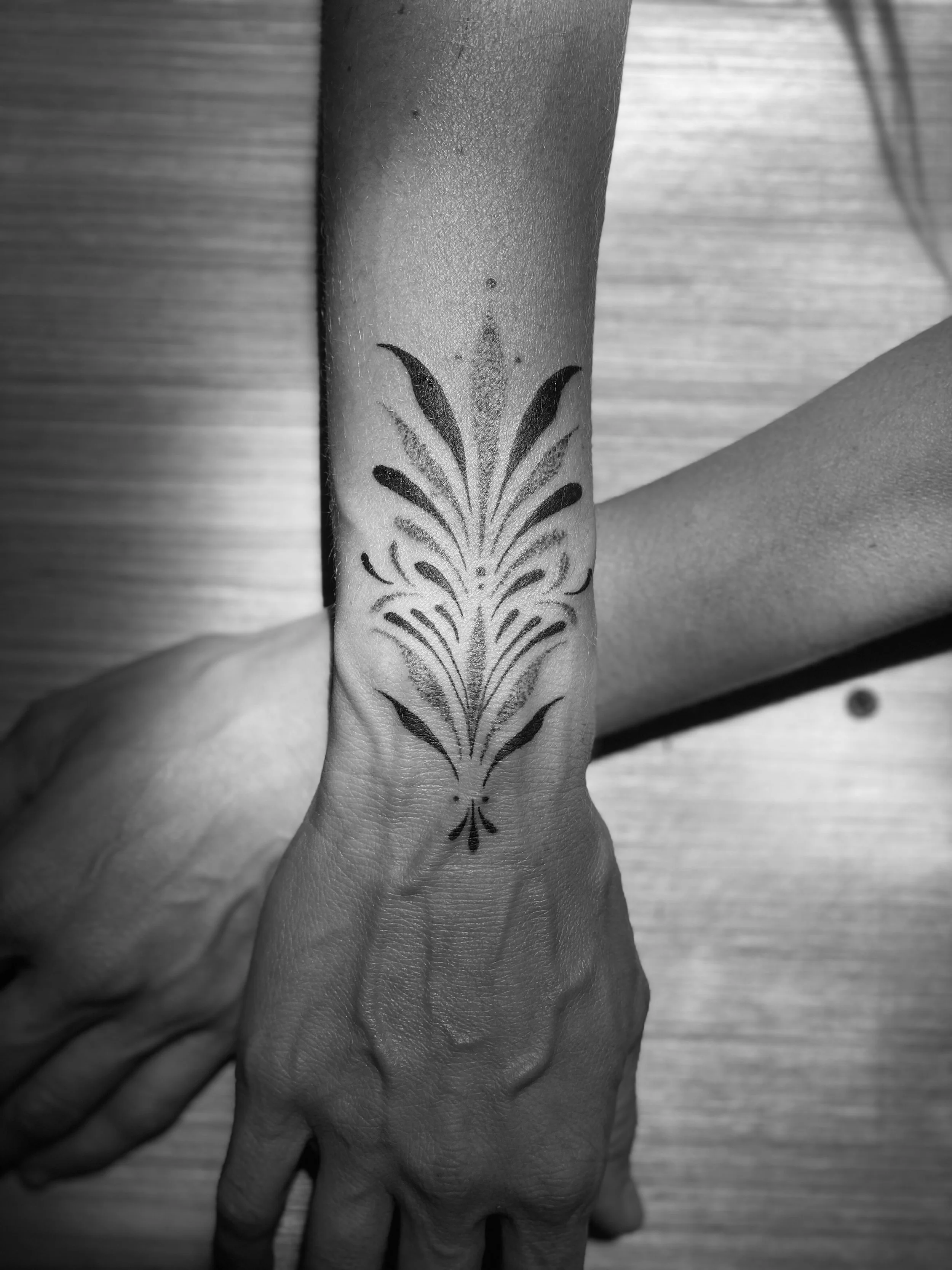 Black and white photo of a person's arm and hand, focusing on a tattoo on the wrist that features symmetrical floral and abstract patterns.