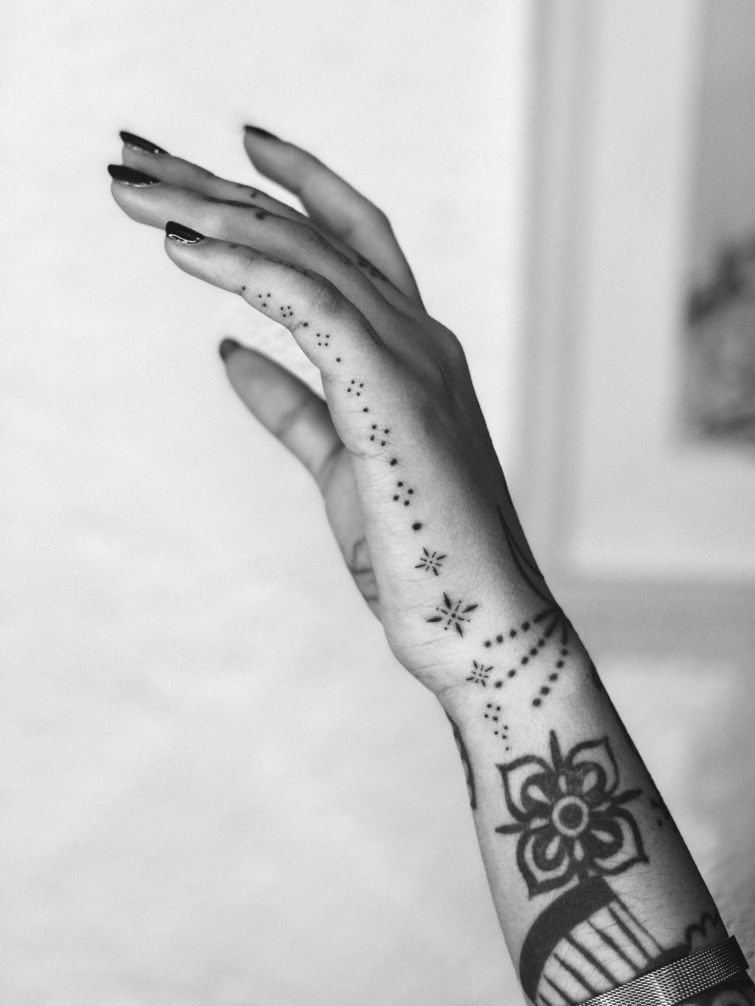 A close-up of a tattooed hand with black nail polish, featuring a floral design and various dotted patterns, against a blurred light background.