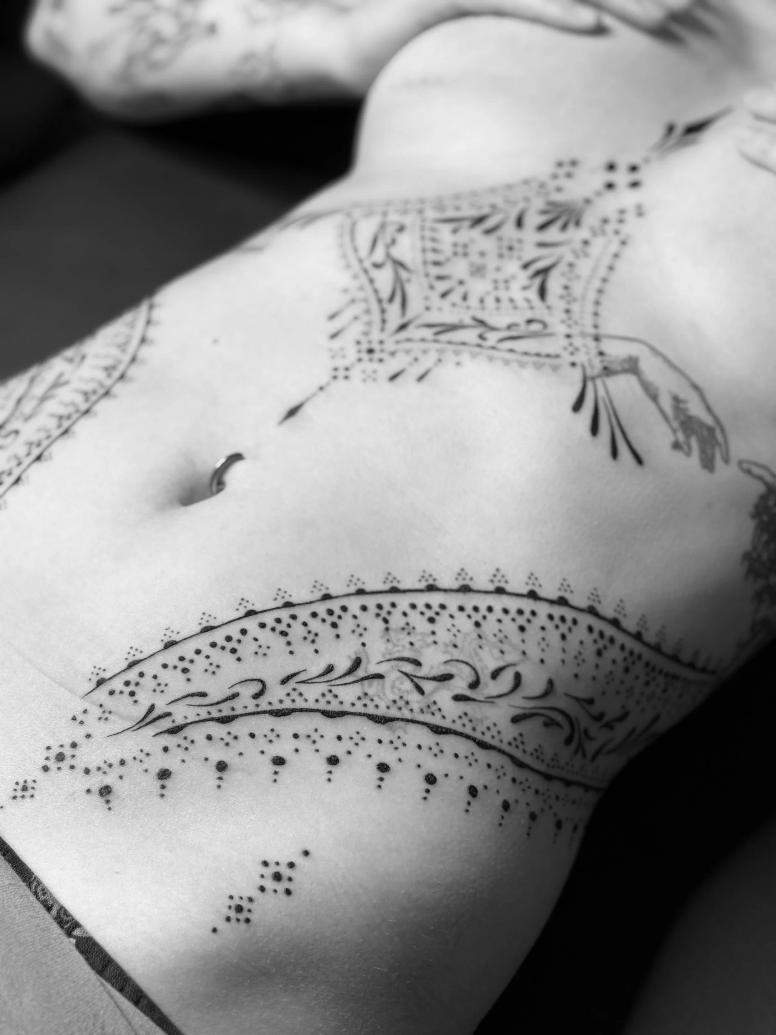 Close-up of a person's torso with tattoo designs and a navel piercing, in black and white.
