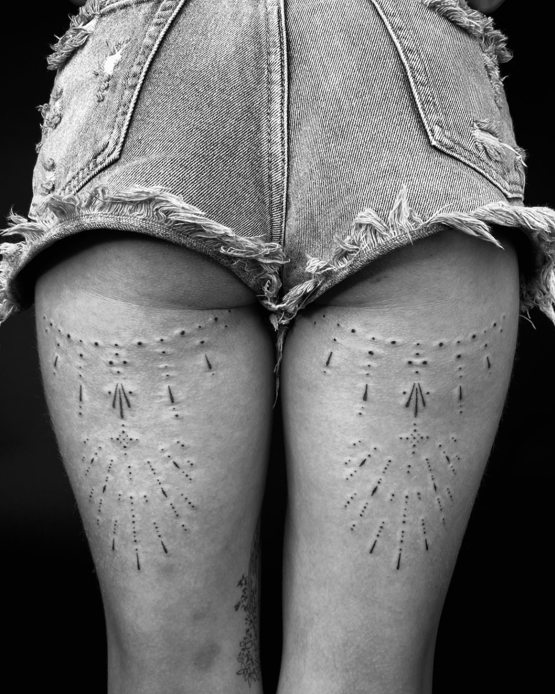 Close-up of a person's backside wearing ripped denim shorts, with tattooed thighs featuring dotted tattoo designs.