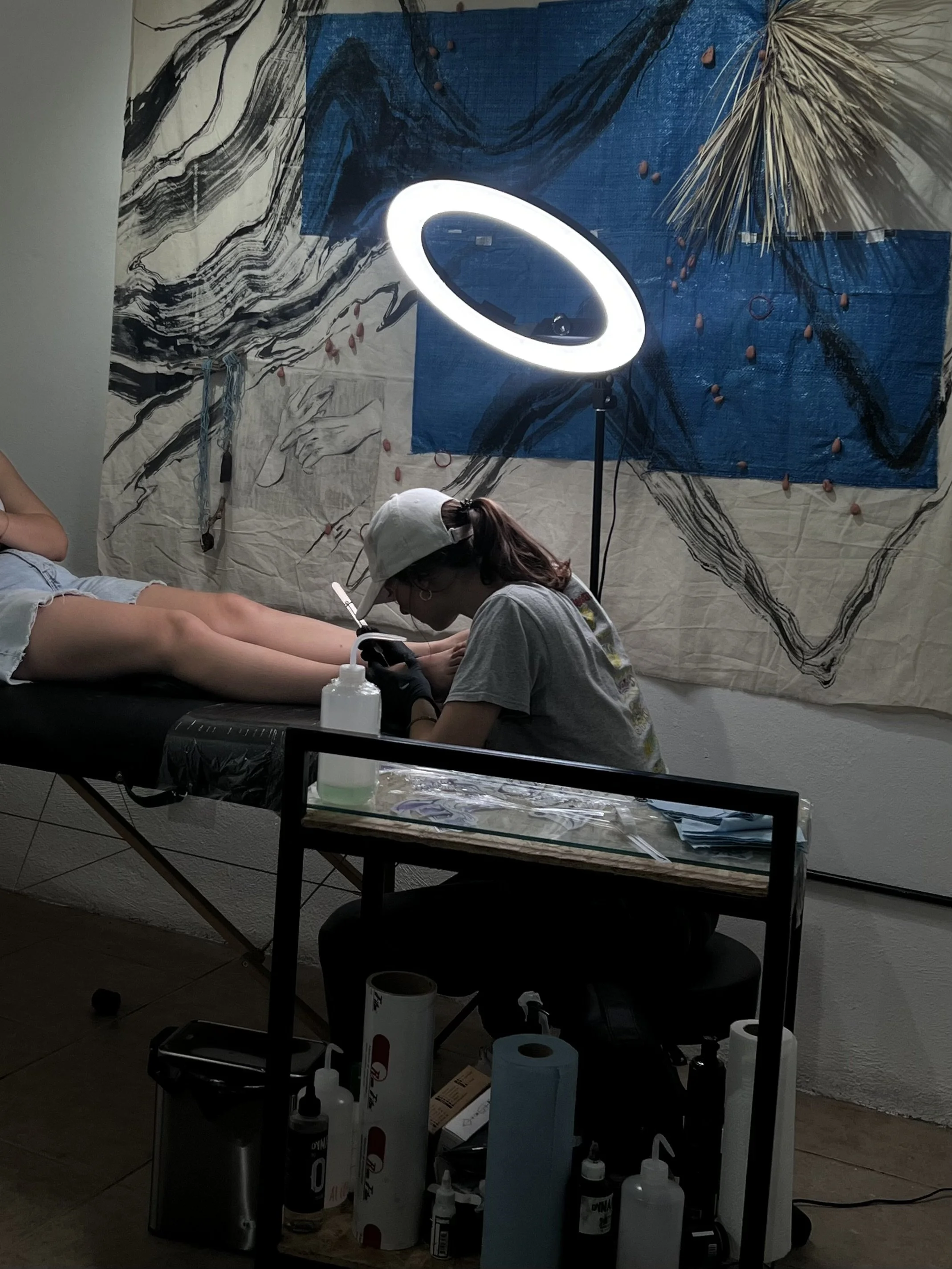 A person receiving a tattoo on their leg in a studio. The artist is focused, wearing gloves and a cap, working under a ring light. The wall behind has abstract art with fabric and hanging decorations.
