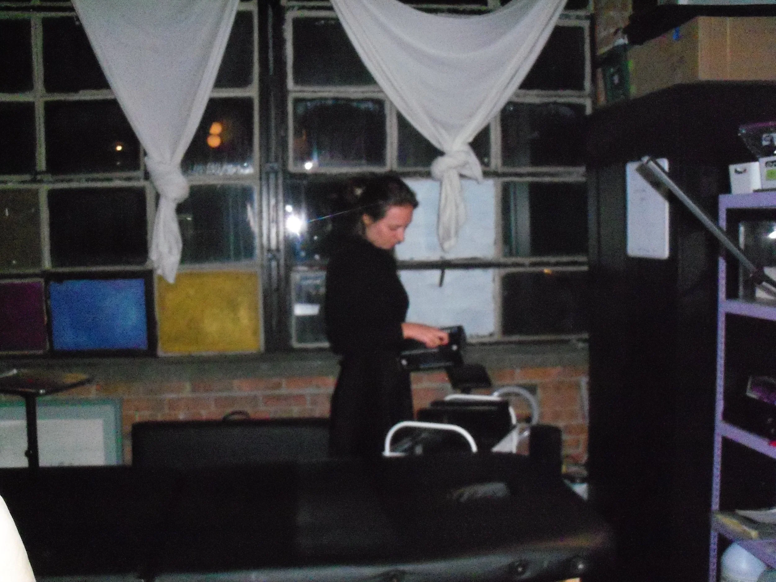 A woman standing indoors near a window with white curtains, looking down at a small object in her hands, with a brick wall and shelves in the background.