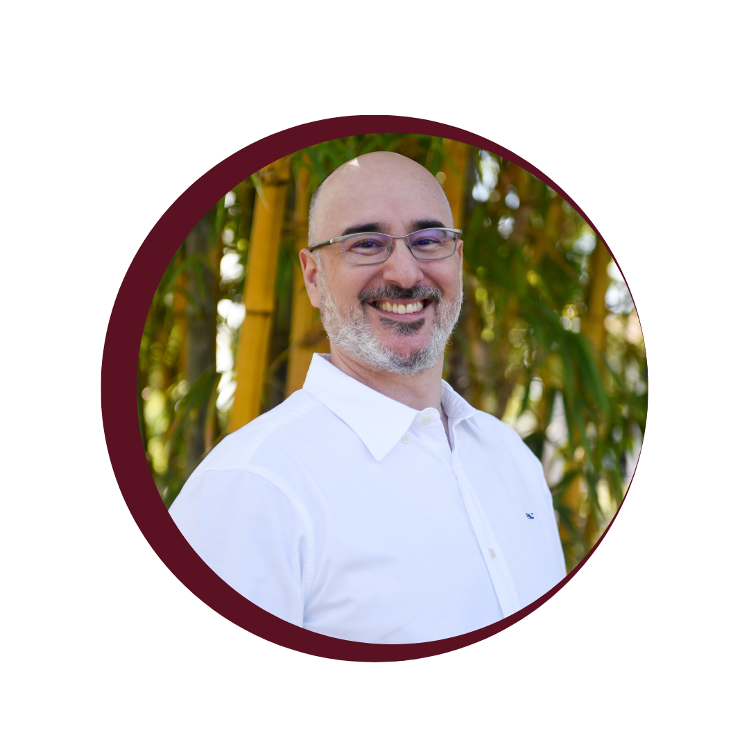 A smiling man with glasses and a beard, wearing a white shirt, standing outdoors among green plants.