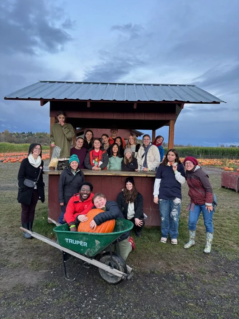 Union Youth Group event with people posing around a pumpkin patch stand at a farm, with pumpkins in the background and a cloudy sky overhead.