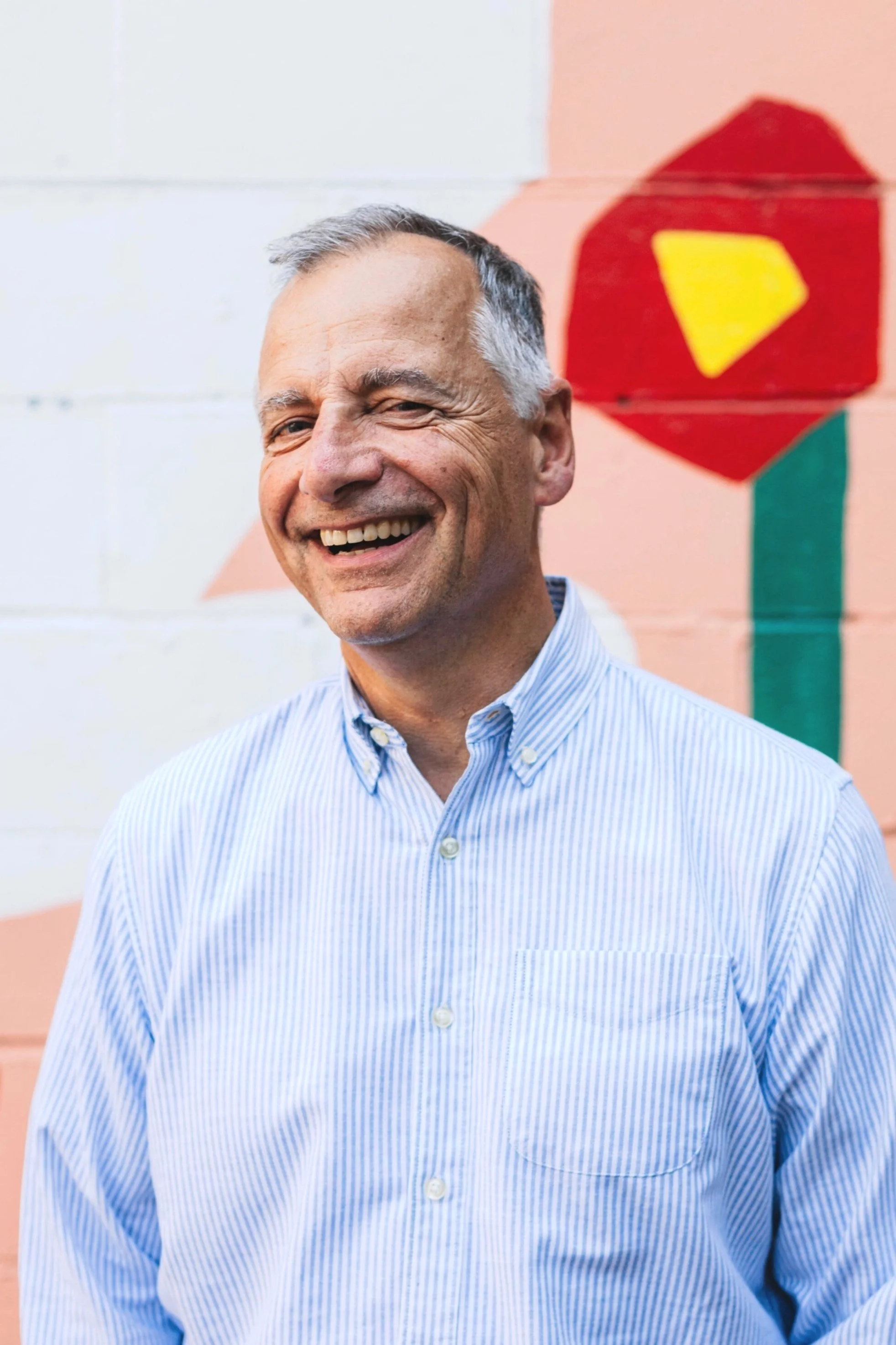 A smiling middle-aged man in a light blue striped shirt stands in front of a painted wall with a colorful flower design.