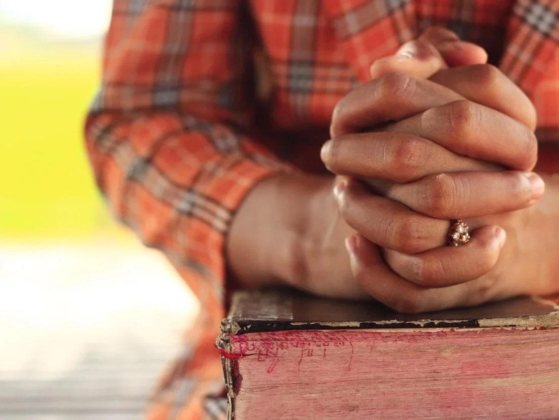 Hands are folded together in prayer, resting on a well used Bible.