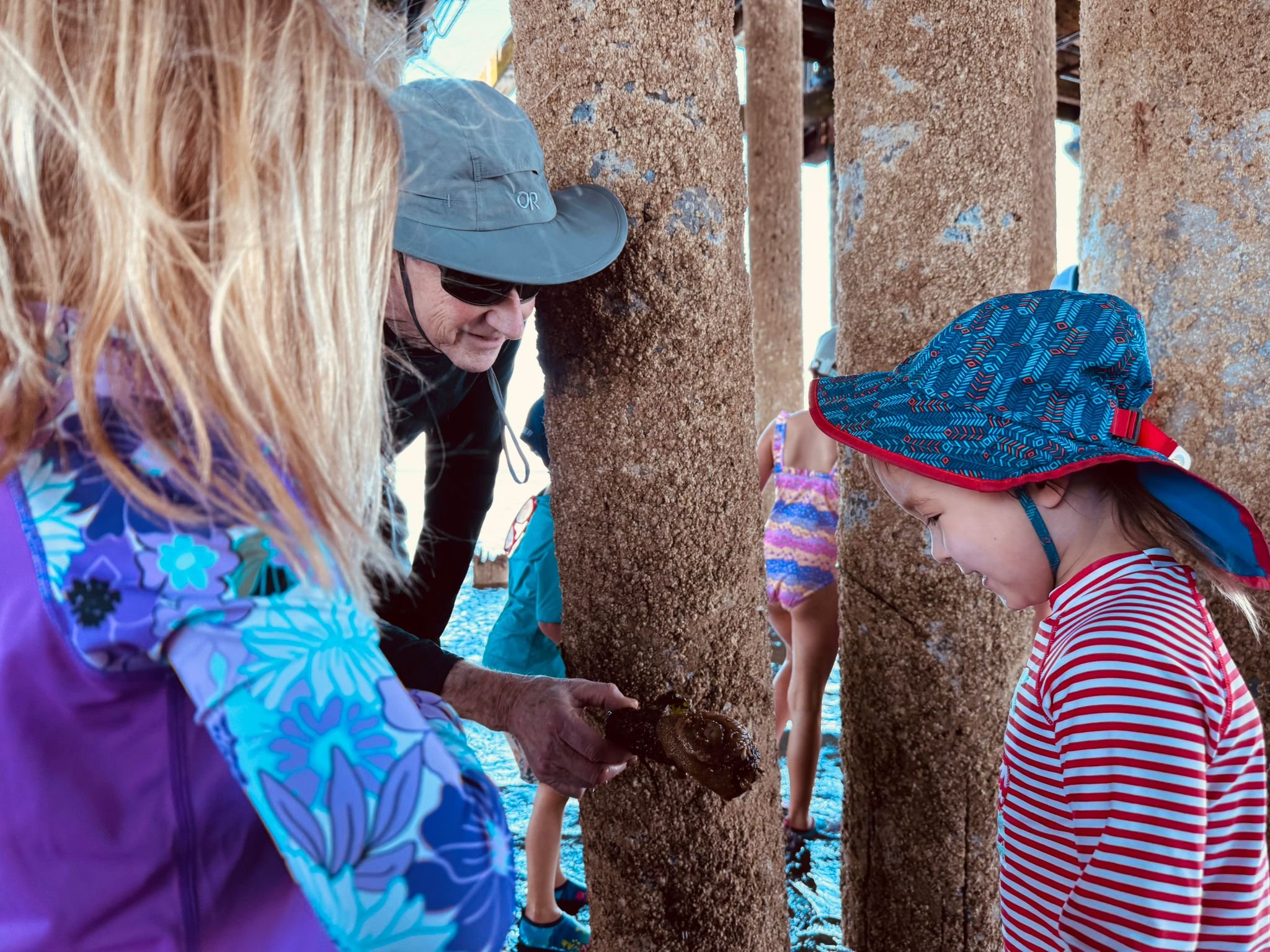 An older man showing a shell to a young girl on a beach with large concrete pillars.