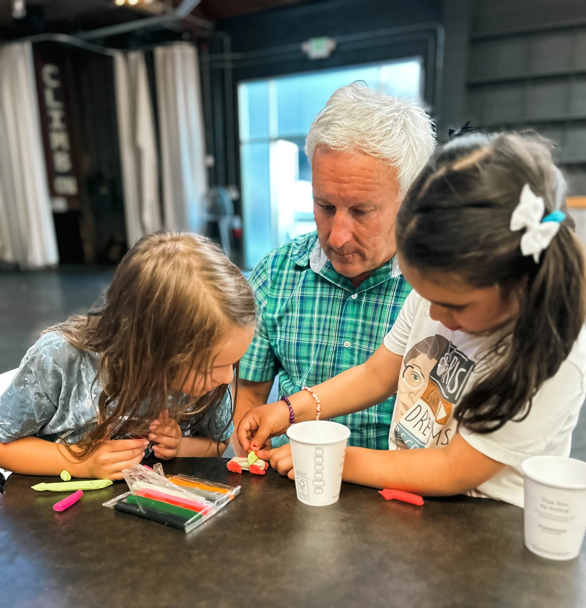 An older man with white hair wearing a green checkered shirt is sitting at a table with two young girls, during our monthly Service Sunday at Union Church in South Lake Union, Seattle.